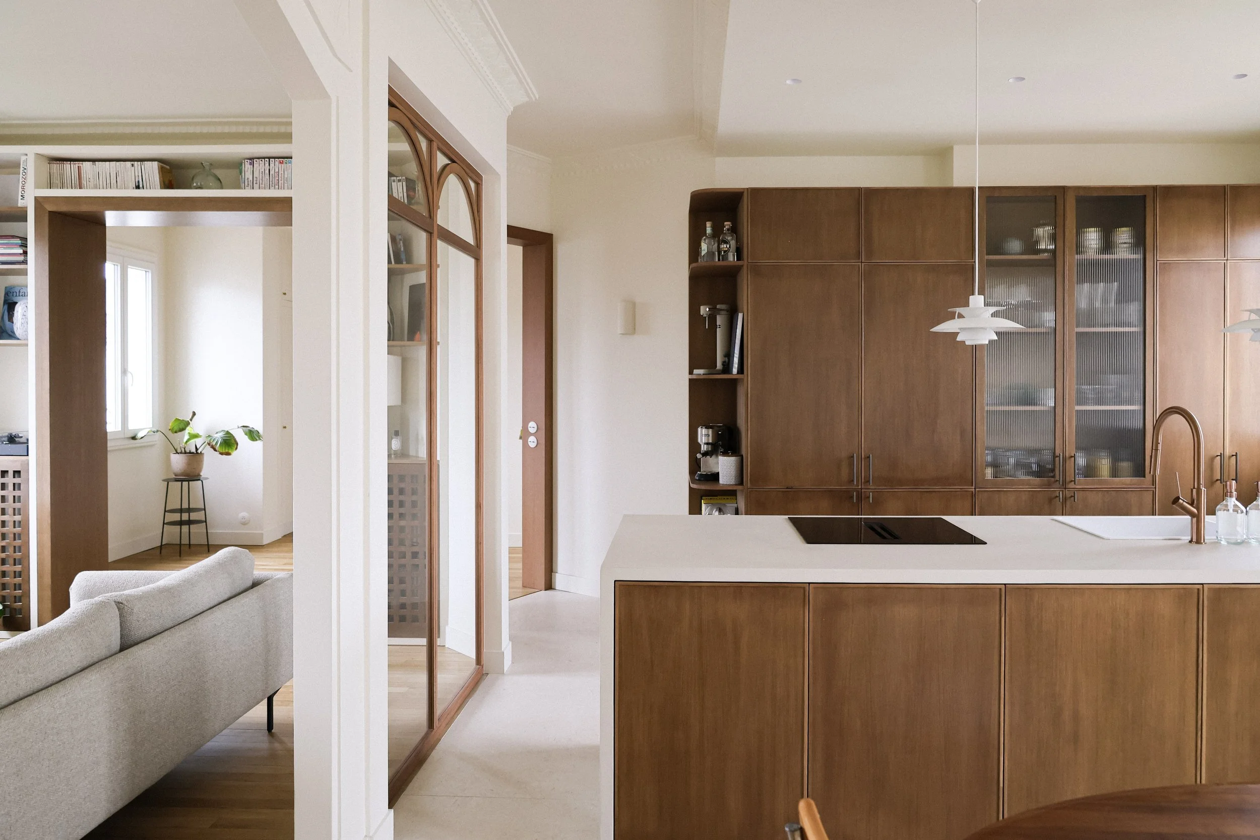 Cuisine ouverte avec îlot central en bois, plan de travail blanc, et armoires en bois. Éclairage suspendu au plafond, évier avec robinet en cuivre, et étagères avec bouteilles et appareils électroménagers. Salon avec canapé gris et étagère avec livre