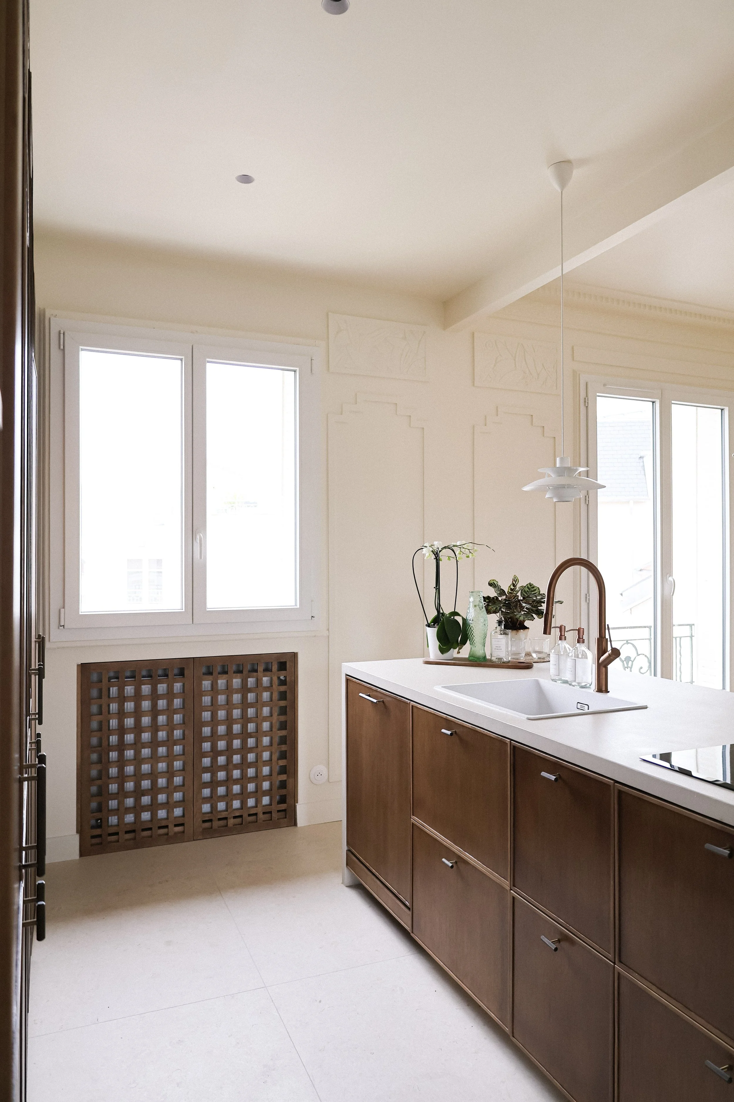 Une cuisine moderne avec un plan de travail blanc, un évier, plusieurs plantes et articles de vaisselle, fenêtres lumineuses, murs écrus et meubles en bois