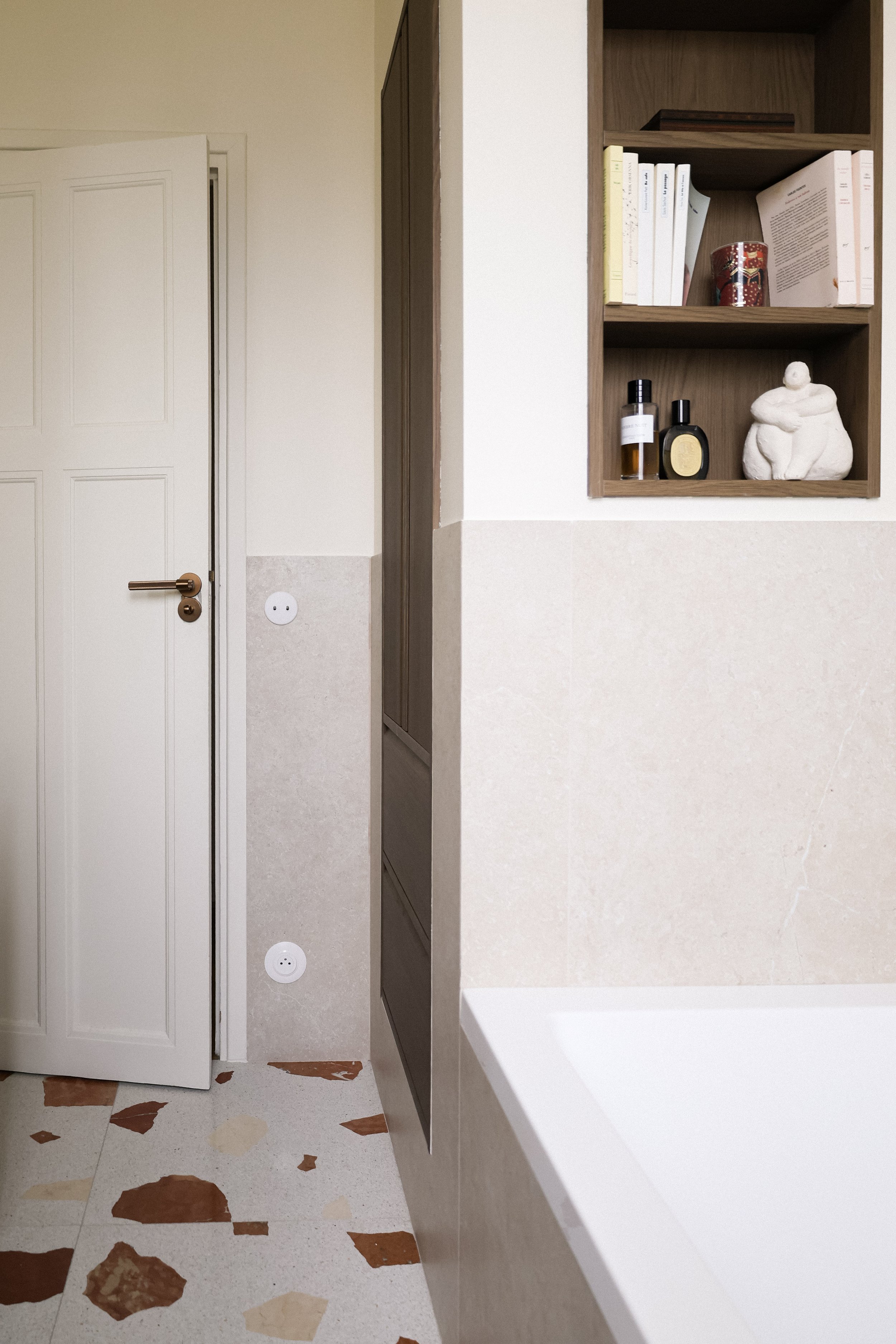 Vue intérieure d'une salle de bain, montrant une porte blanche, une étagère en bois avec des livres, des bouteilles de parfum, et une sculpture abstraite, avec un mur beige et un sol en carreaux semi-circulaires.