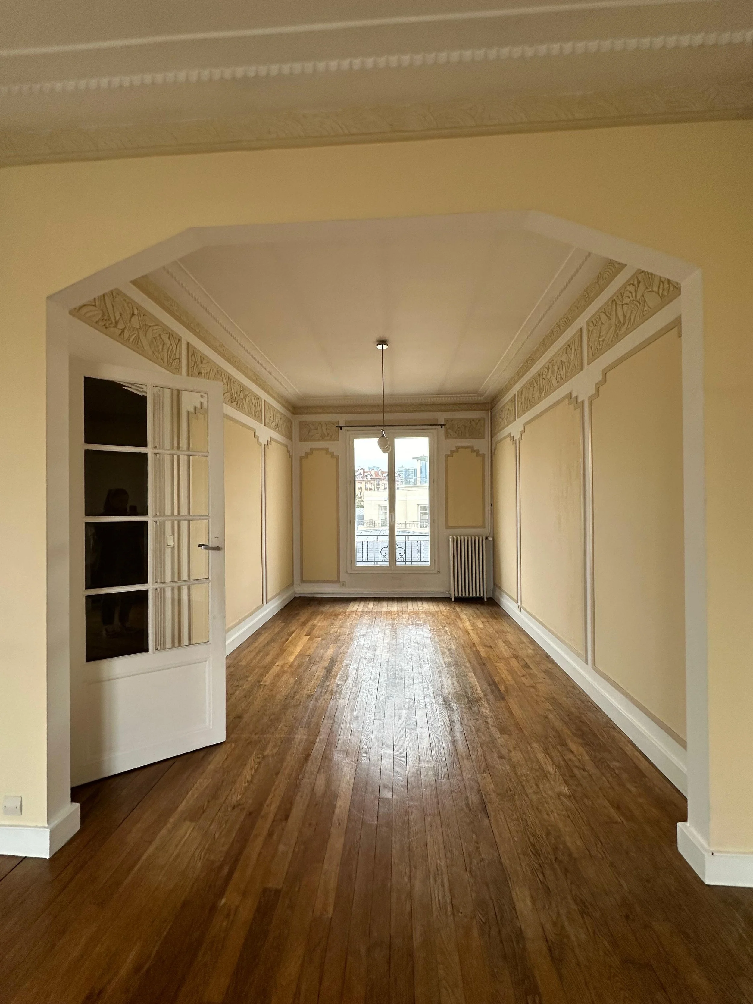 Une pièce vide avec des murs jaune clair, des moulures ornementales et un parquet en bois clair, une fenêtre avec un radiateur en dessous et une porte en verre ouvrant sur une autre pièce.