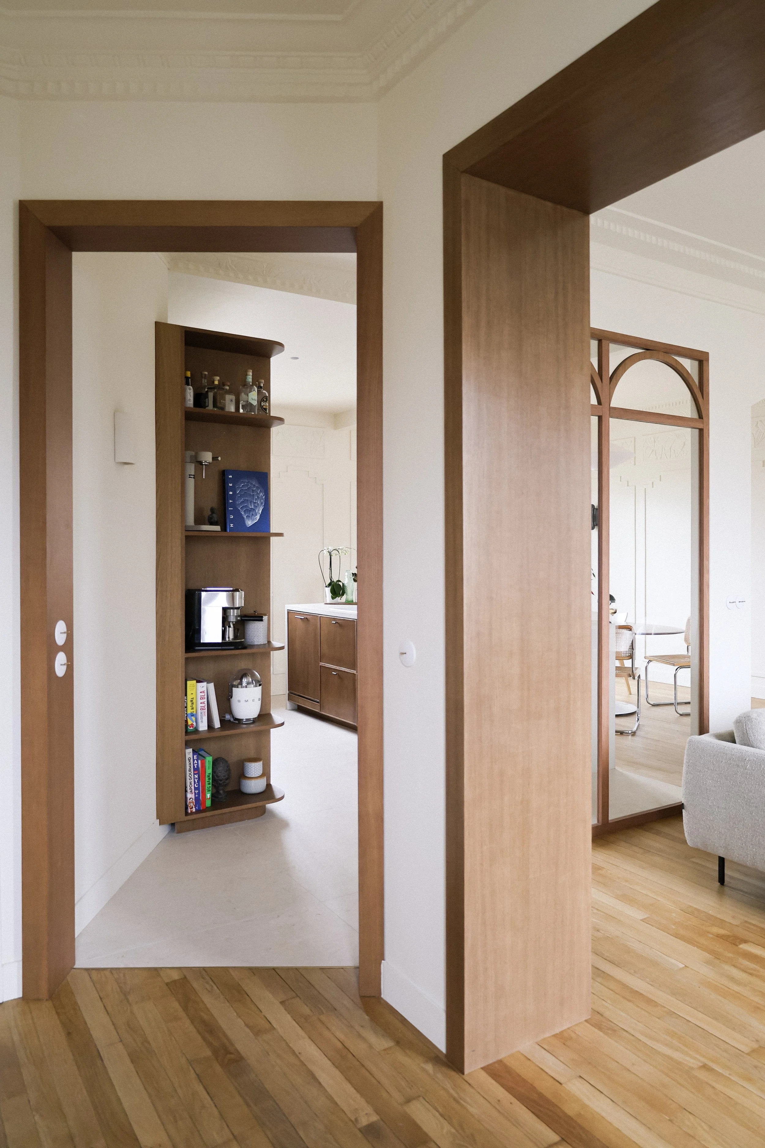 Intérieur moderne d'une maison avec portes en bois, vue sur une étagère avec des livres et des objets, et un salon avec un canapé beige.