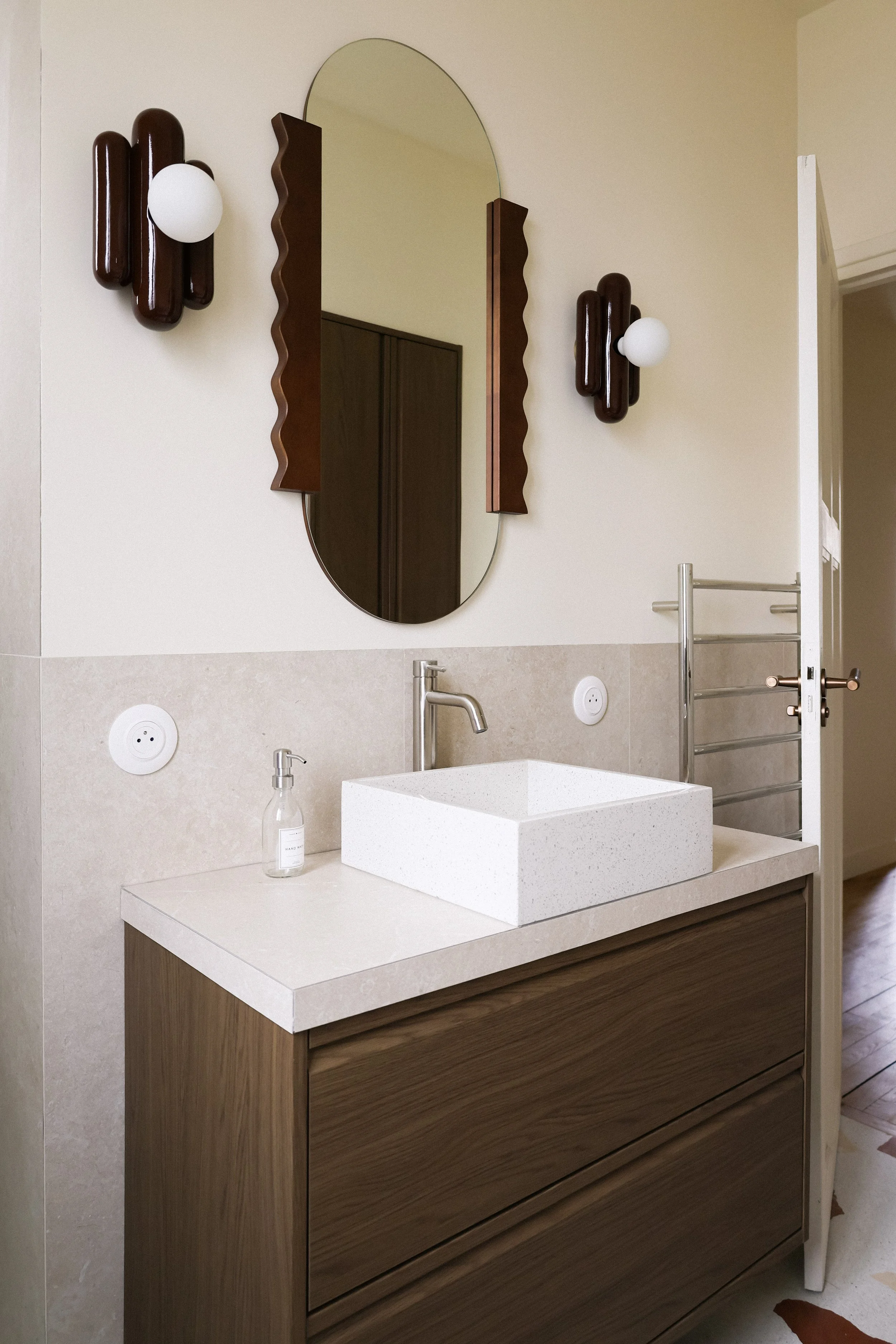 Intérieur d'une salle de bain moderne avec un lavabo en forme de cube en pierre, un miroir ovale avec un cadre en bois sculpté, deux appliques murales en bois foncé avec des globes blancs, un distributeur de savon, un sèche-serviettes en métal, et un