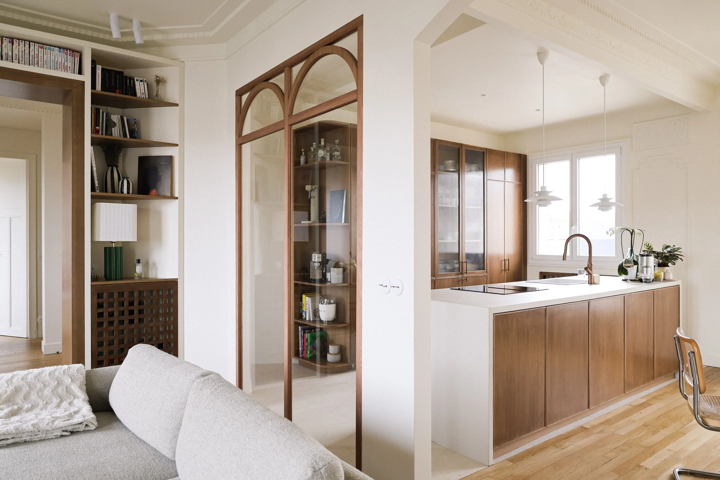 Intérieur d'une cuisine moderne avec armoires en bois, comptoir blanc, fenêtres laissant entrer la lumière naturelle, quelques plantes vertes sur le comptoir, et un coin salon avec un canapé beige et une couverture blanche
