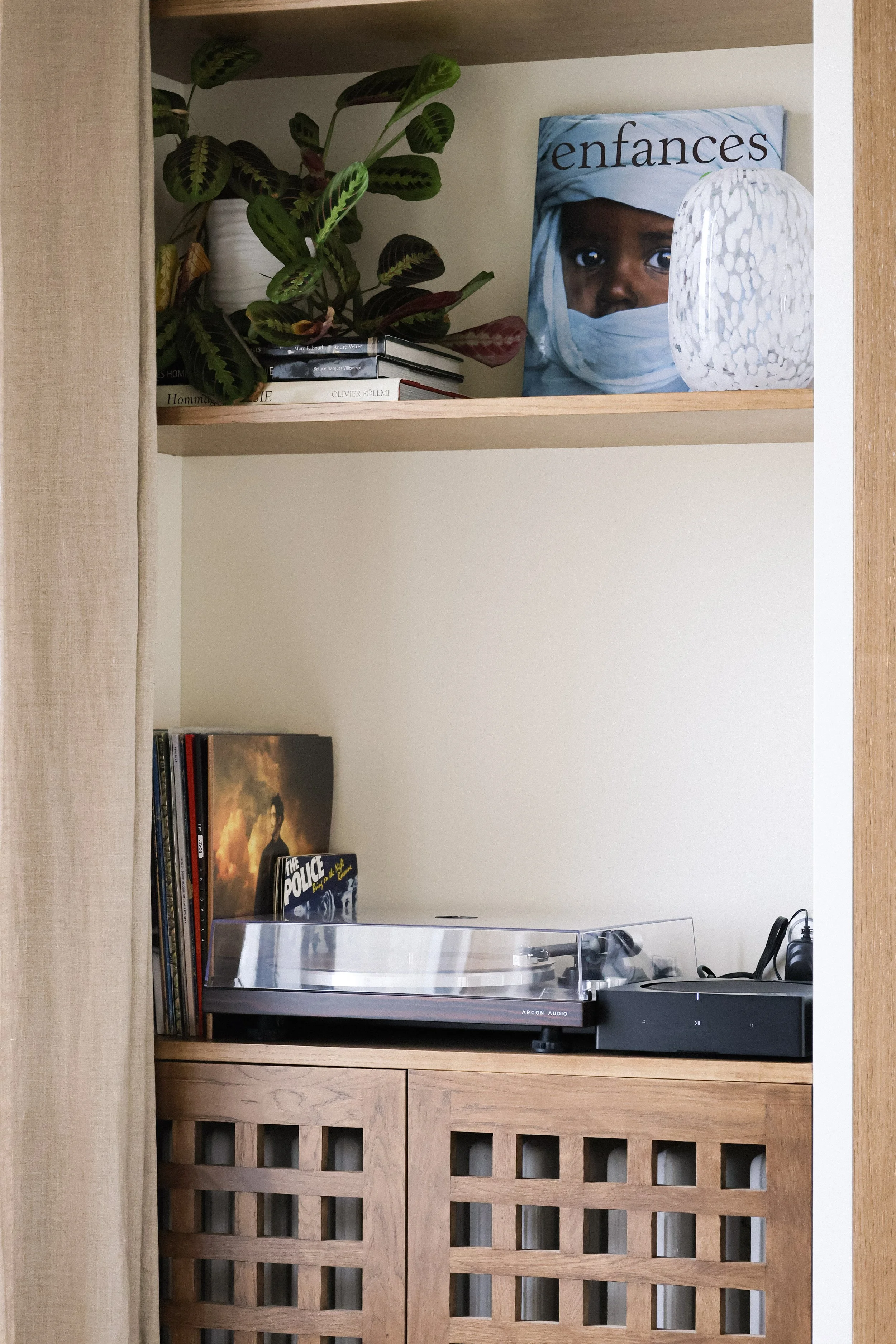 Une étagère avec des livres, une plante verte en pot, une photo d'enfant en couverture d'un magazine, une lampe blanche, un tourne-disque et une enceinte audio, avec un meuble en bois en dessous.