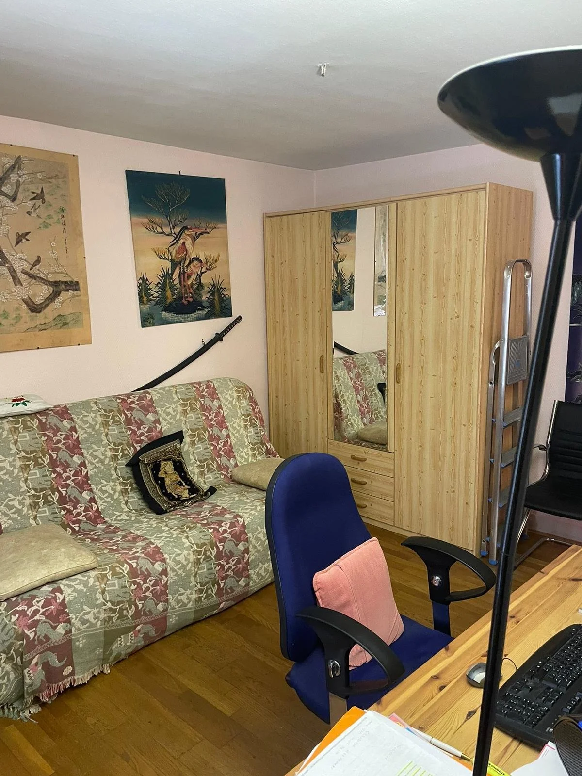Un salon avec un canapé recouvert d'une housse à motifs, un fauteuil de bureau bleu avec un coussin rose, un mur avec des œuvres d'art japonais, une armoire en bois avec des miroirs, un escabeau en aluminium, un ordinateur et une lampe sur un bureau.
