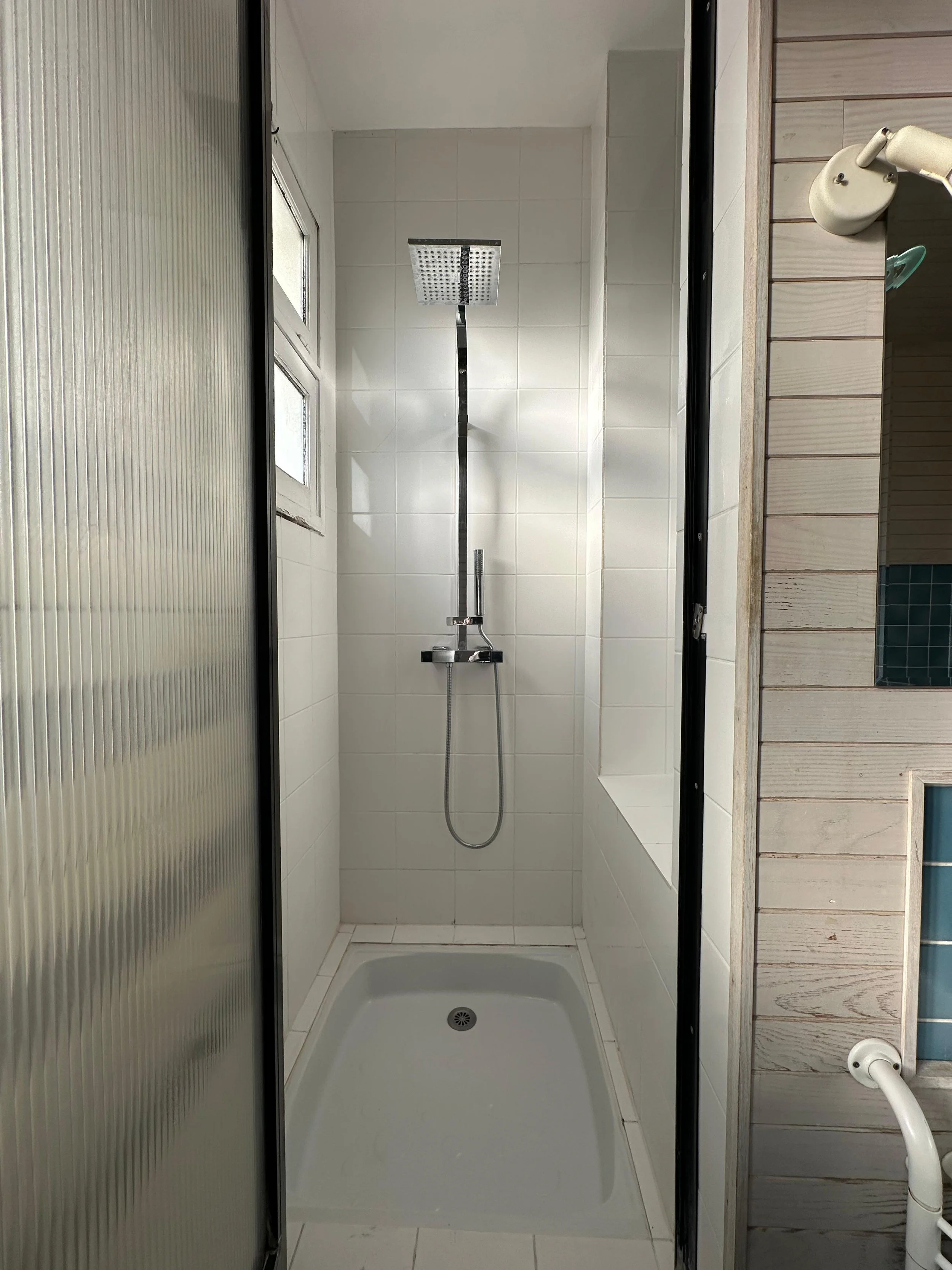 Douche dans une cabine avec mur en carrelage blanc, pomme de douche carré suspendue, parois en verre texturé, et fenêtre en haut à gauche.