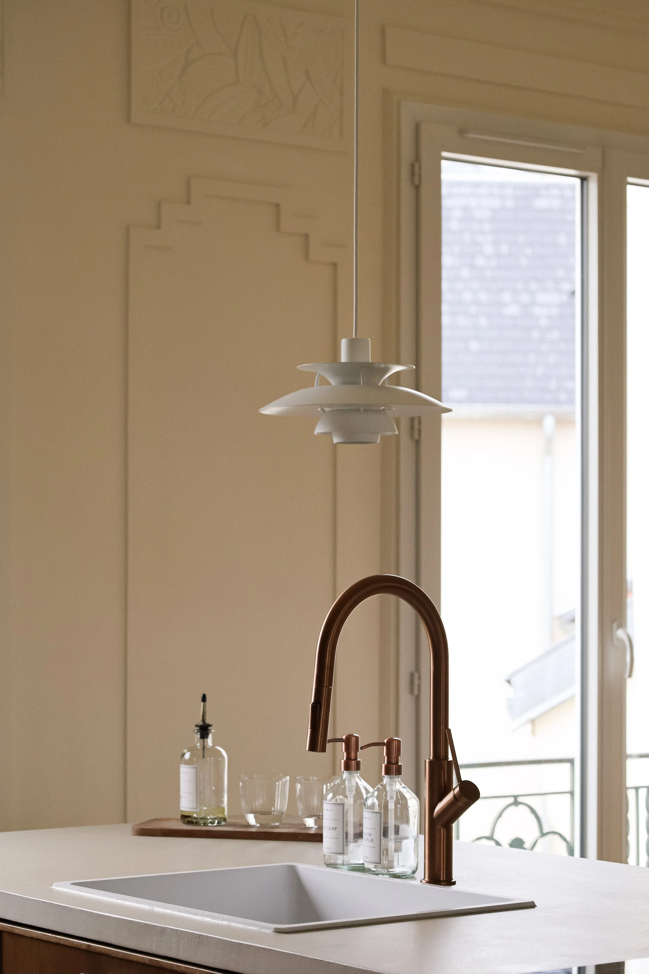 Évier de cuisine avec robinet en cuivre, bouteilles de savon à mains, et grande fenêtre laissant entrer la lumière naturelle.