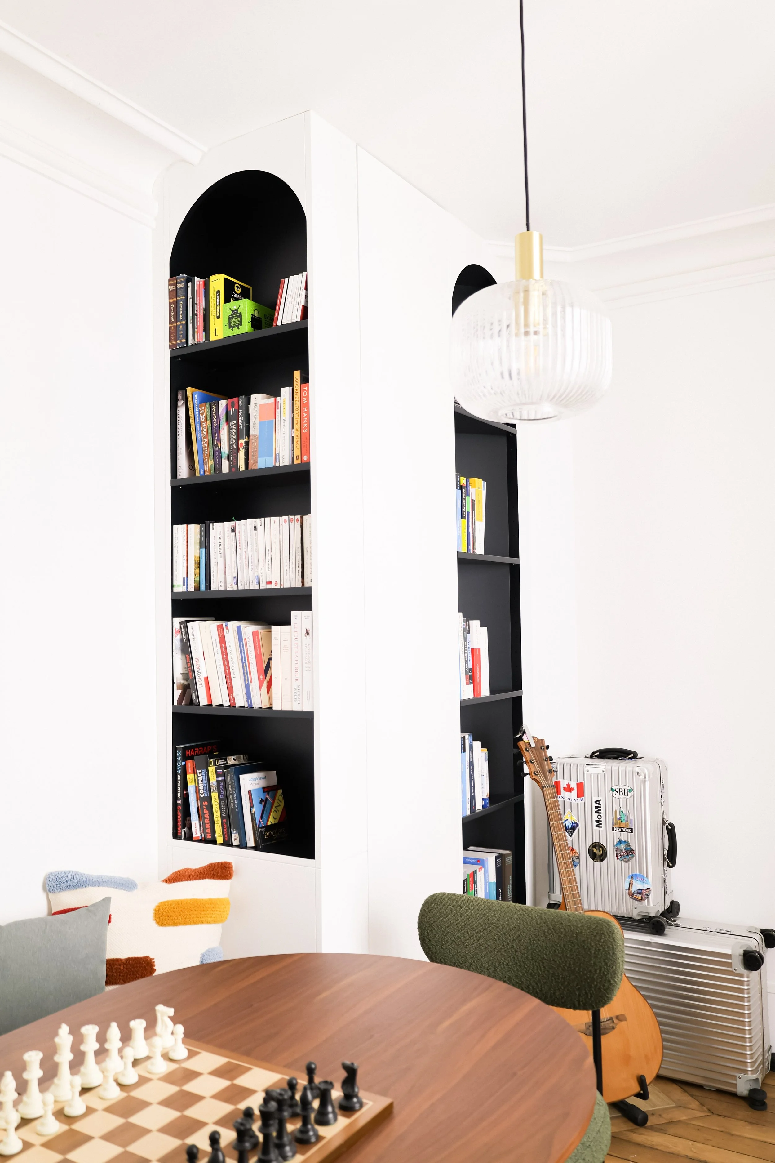 Une salle de séjour avec une grande table en bois, une chaise verte, une étagère noire remplie de livres, une guitare, deux valises, un luminaire suspendu blanc, des coussins colorés, un parquet en bois, et un mur blanc.