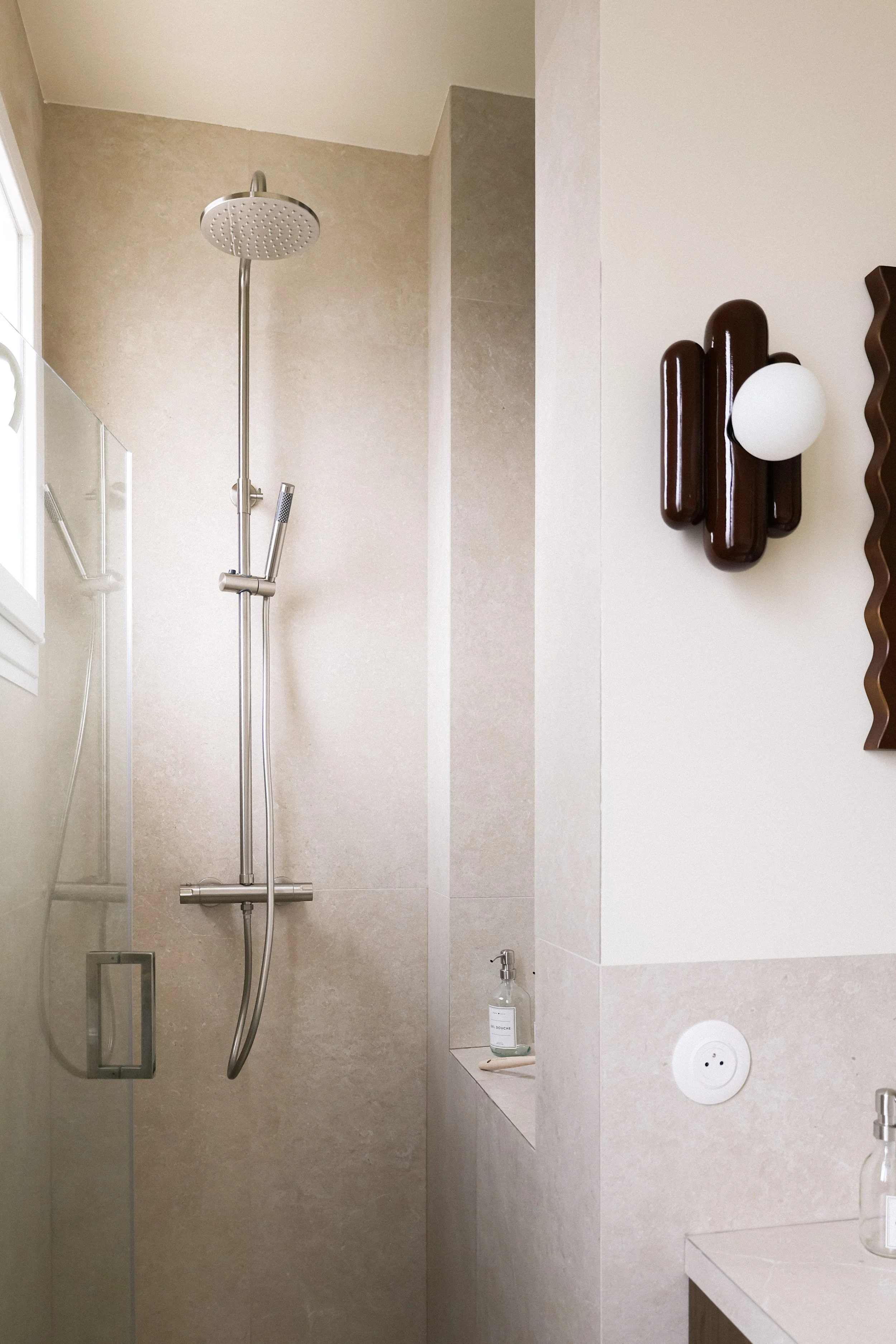 Salle de bain moderne avec douche à l'italienne, mur beige, prise électrique ronde blanche, miroir moulé wavy, bouteille de savon liquide, et décoration murale en bois.