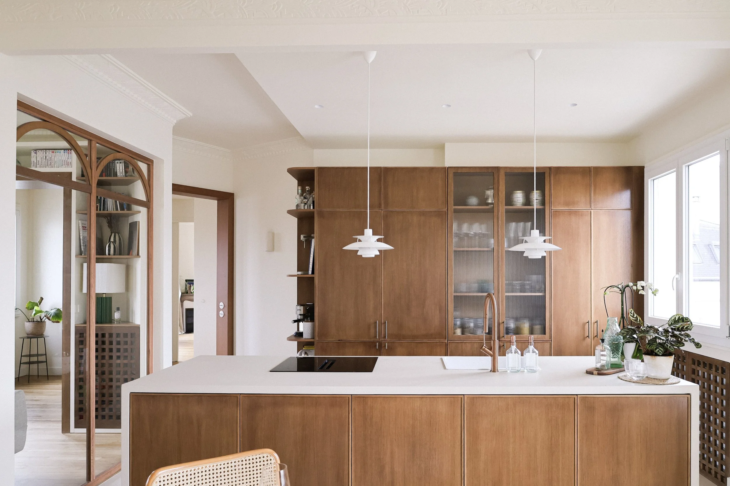 Cuisine moderne avec îlot central en pierre, armoires en bois, suspensions blanches, plantes vertes près de la fenêtre, espace lumineux.