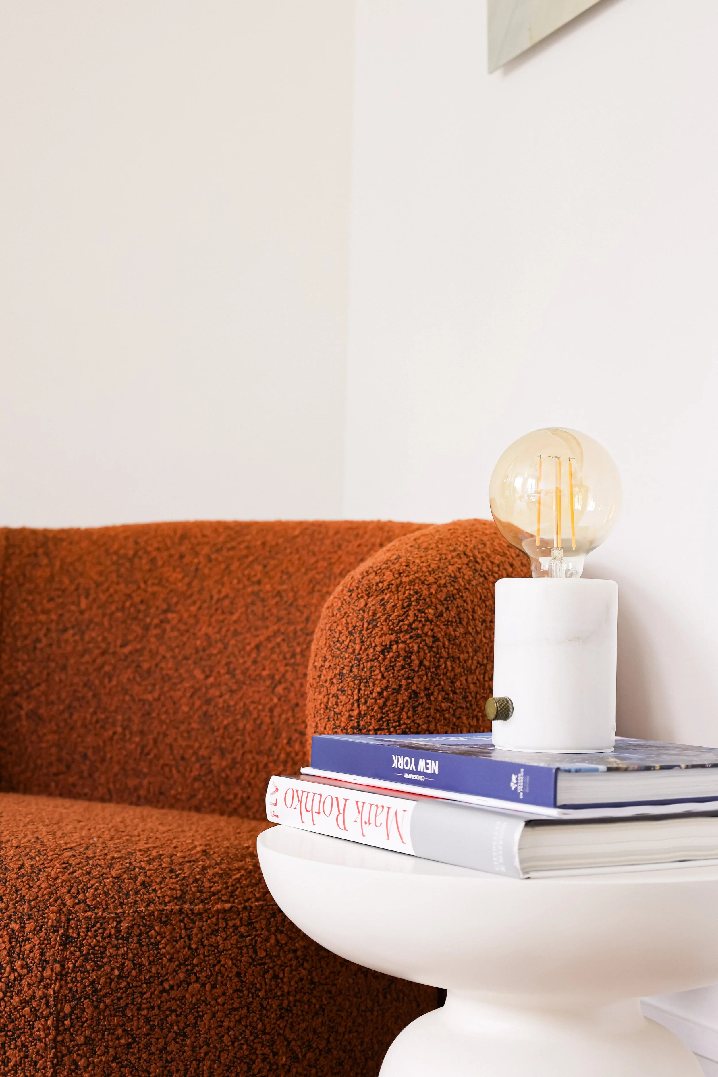 Table avec livres et lampe à ampoule visible, canapé orange en arrière-plan, mur blanc.