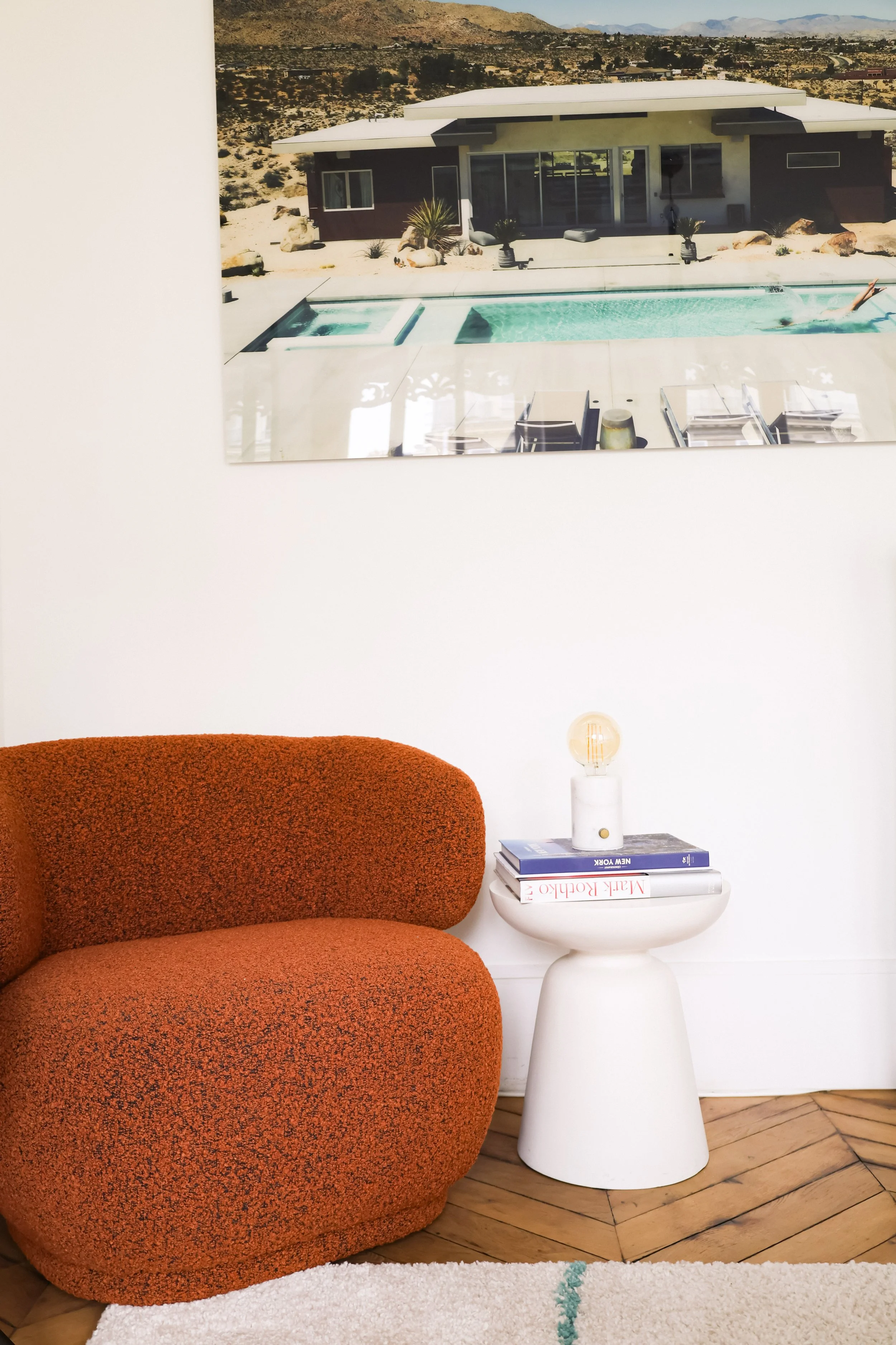 Intérieur moderne avec un fauteuil orange textured, une petite table blanche avec des livres et une ampoule décorative, et un grand tableau représentant une maison avec piscine à l'extérieur, dans un environnement désertique.