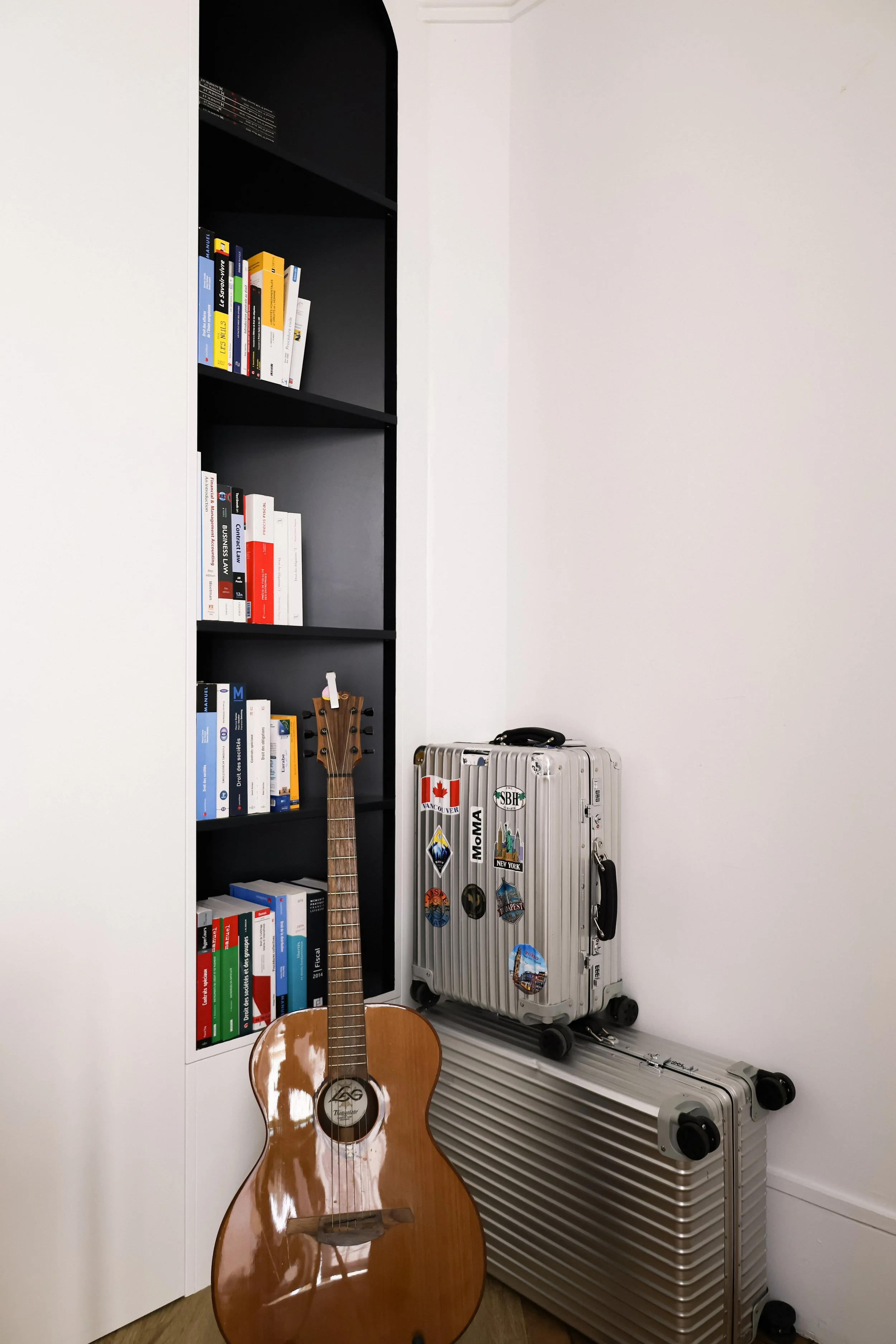Une étagère noire avec des livres, une guitare acoustique, deux valises en aluminium, une blanche avec des autocollants et une grise