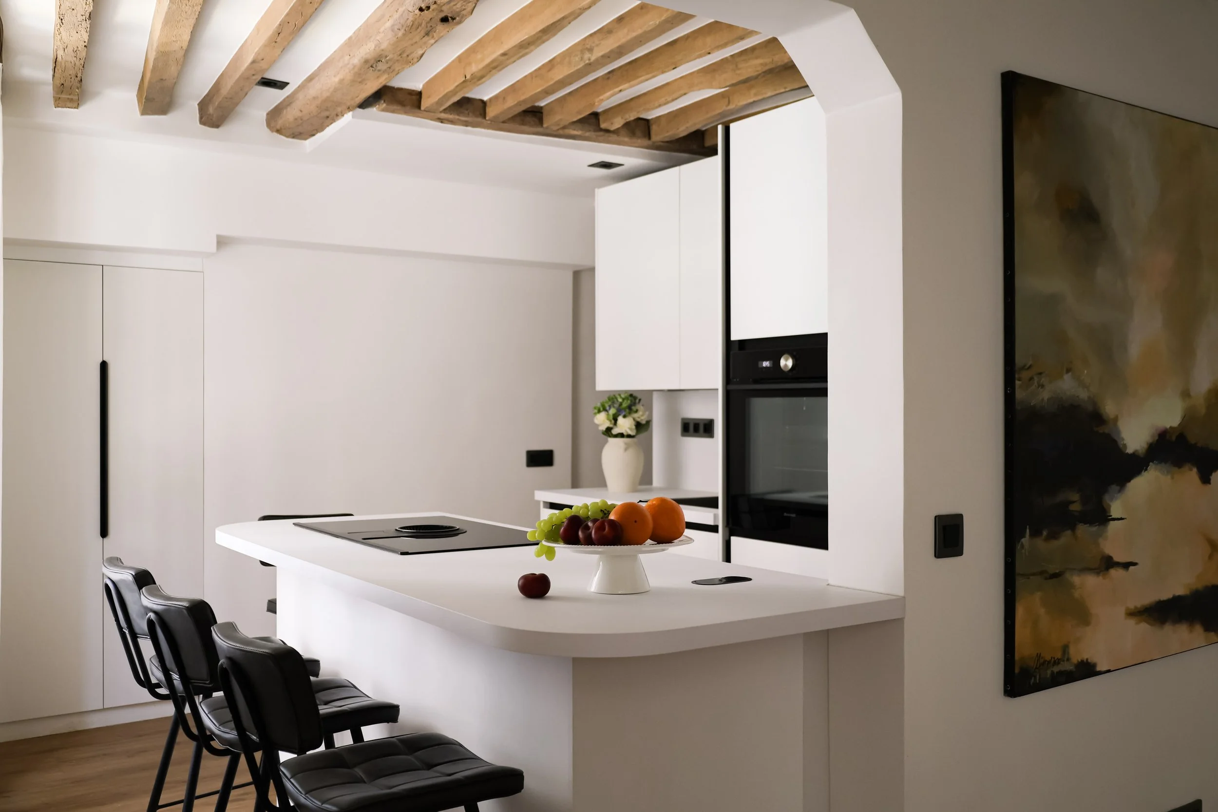 Cuisine moderne avec un plan de travail blanc, des chaises noires, un four intégré, un vase avec des fleurs, et un tableau abstrait.