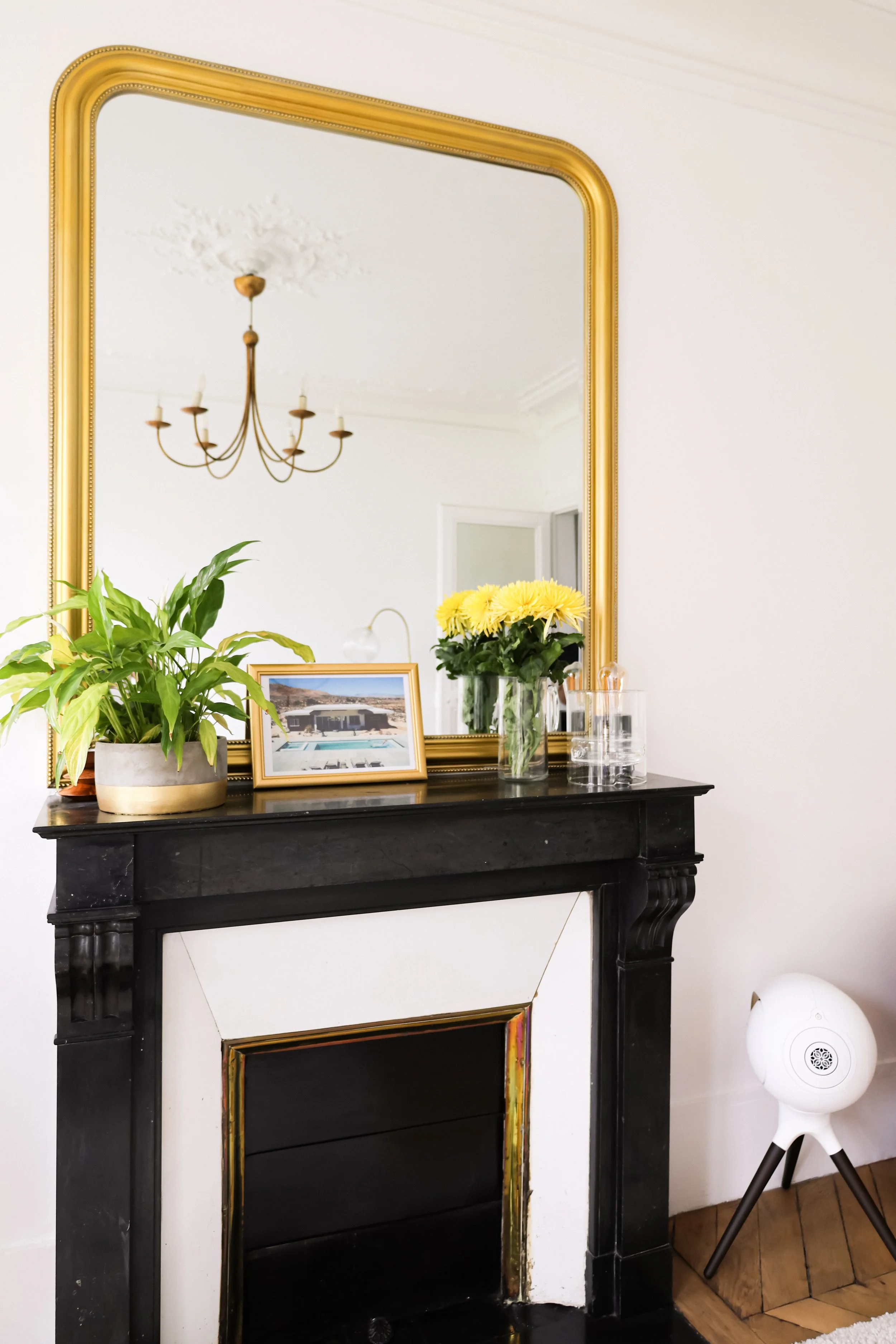 Un miroir doré au-dessus d'une cheminée noire avec des décorations, une plante verte en pot, un vase avec des fleurs jaunes, un cadre photo, des verres en verre, une lampe de chevet, et un appareil de diffusion d'air blanc sur un sol en bois.