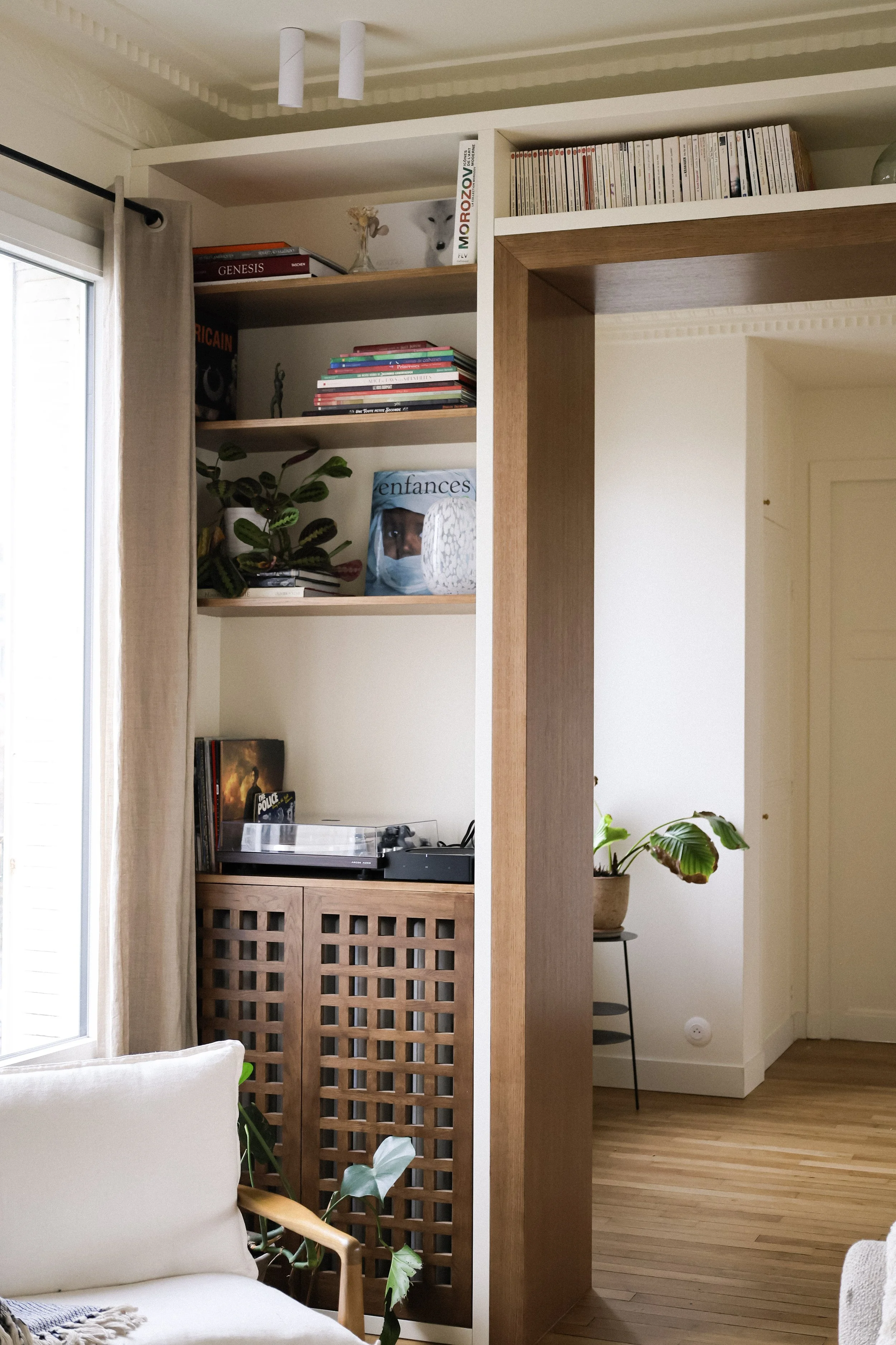 Vue intérieure d'un salon avec étagères en bois contenant des livres, des plantes et des objets décoratifs, un tourne-disque sur un meuble en bois, un canapé blanc avec coussin, et une fenêtre avec rideau beige.