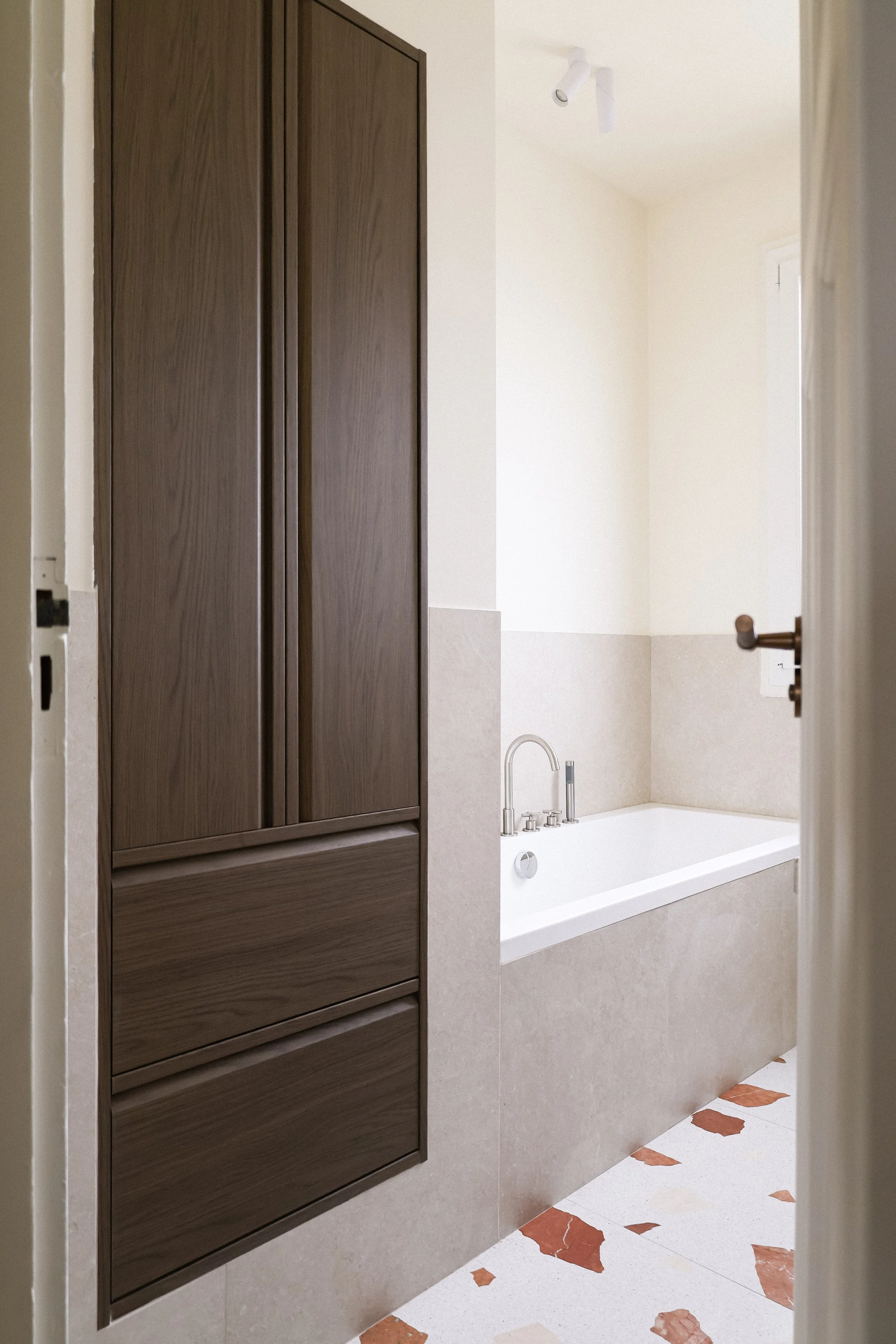 Vue partielle d'une salle de bain avec une baignoire blanche, un robinetterie en acier inoxydable, un mur beige clair, et un placard en bois foncé.