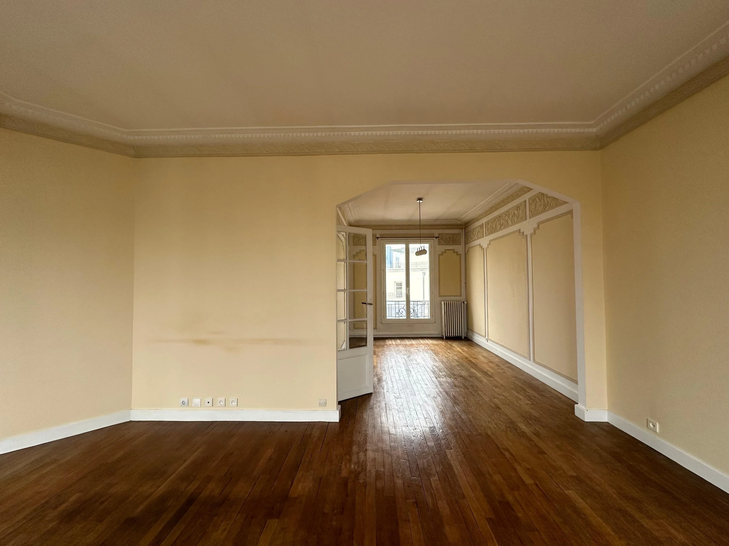 Un grand salon avec des murs jaunes, un plafond orné de moulures, un parquet en bois foncé et une grande fenêtre donnant sur une vue urbaine.