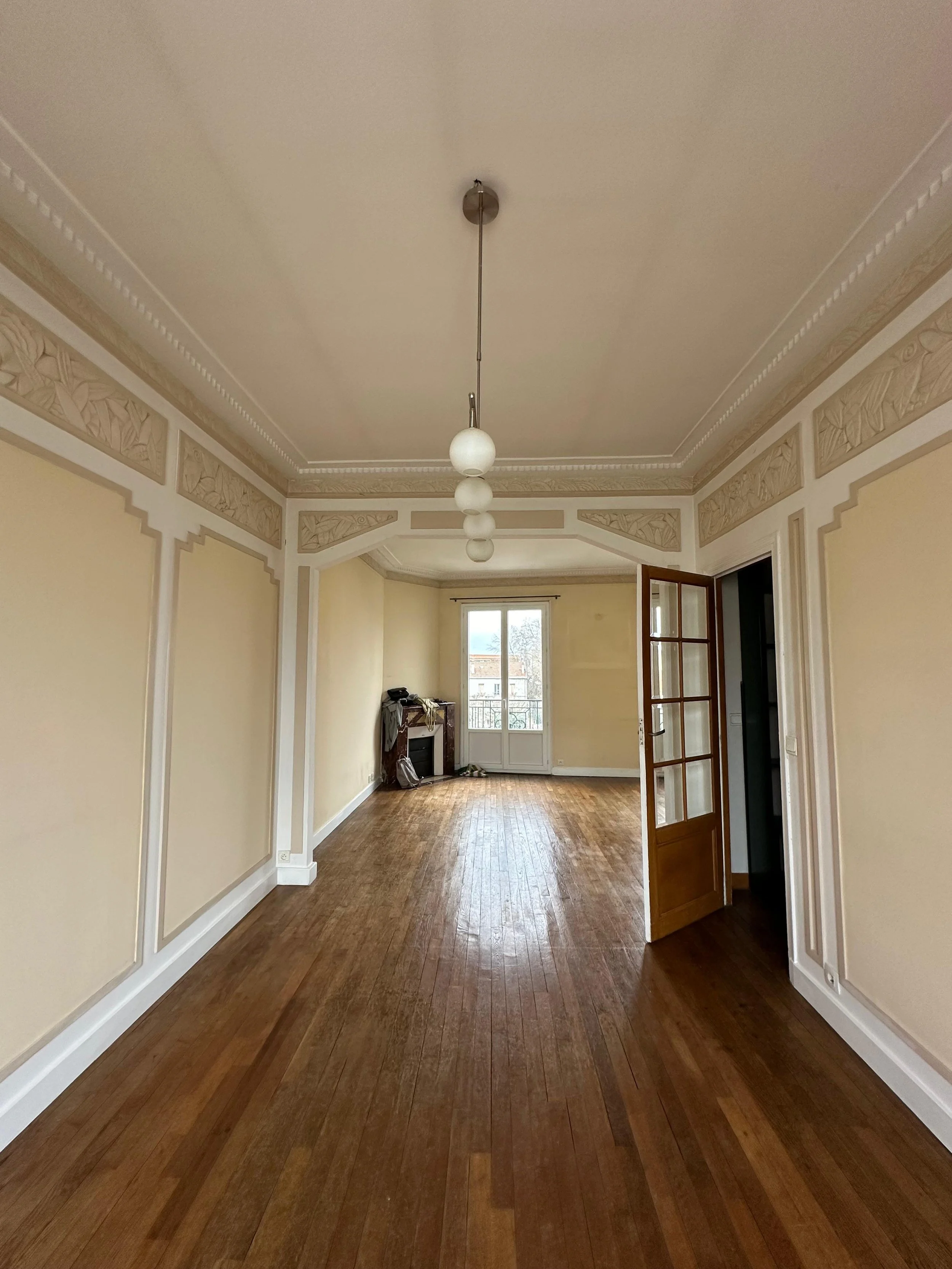Grand salon vide avec parquet en bois, murs crème avec moulures décoratives, plafond à lampes suspendues, porte en bois vitrée, et une grande fenêtre donnant sur un balcon avec vue sur le quartier.