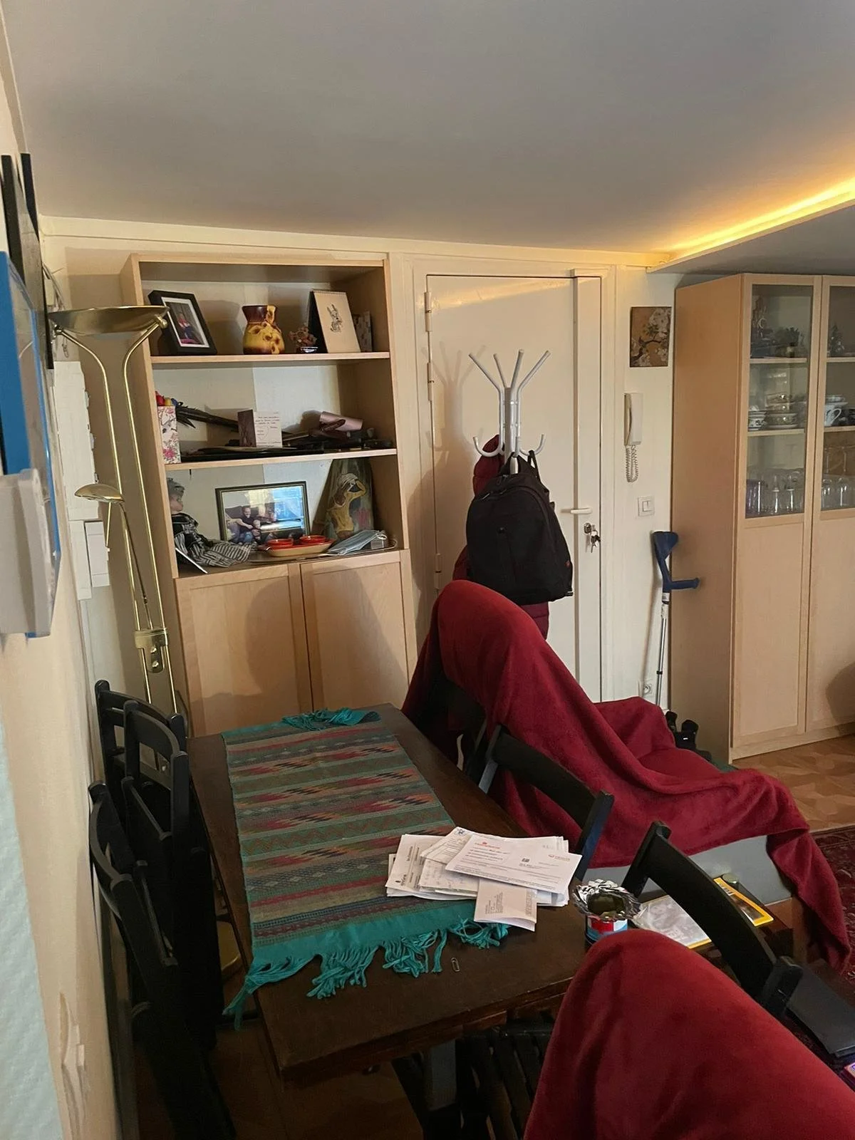 Un salon avec une table en bois recouverte d'une nappe colorée, des chaises autour, un fauteuil rouge couvert d'un tissu rouge, une étagère avec des cadres et des objets décoratifs, un porte-manteau avec des sacs, et des meubles de rangement en bois.