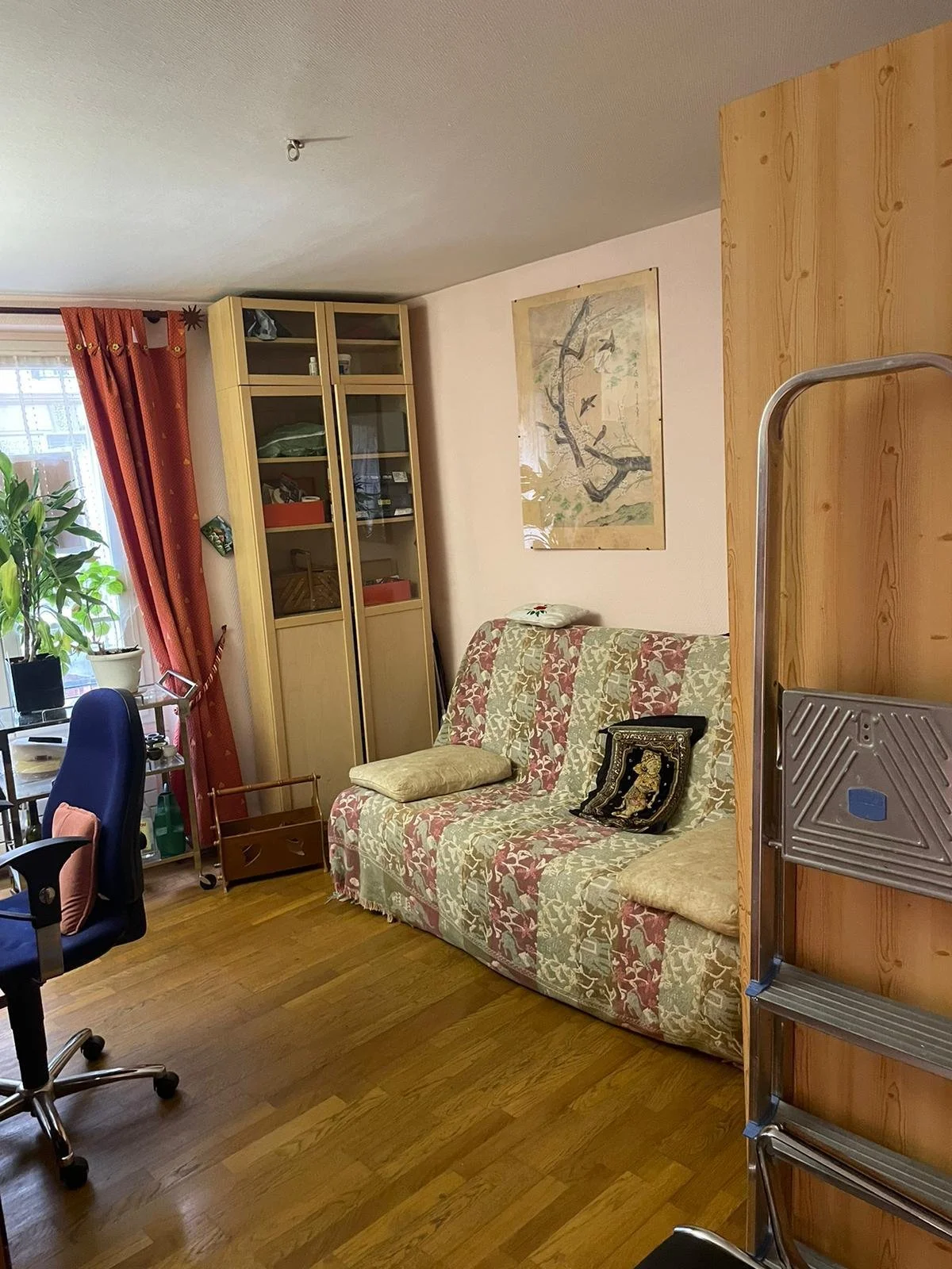 Salon avec canapé à motifs, étagère en bois, table près de la fenêtre, rideaux rouges, plante verte, tableau d'oiseaux, chaise de bureau, escabeau en métal.