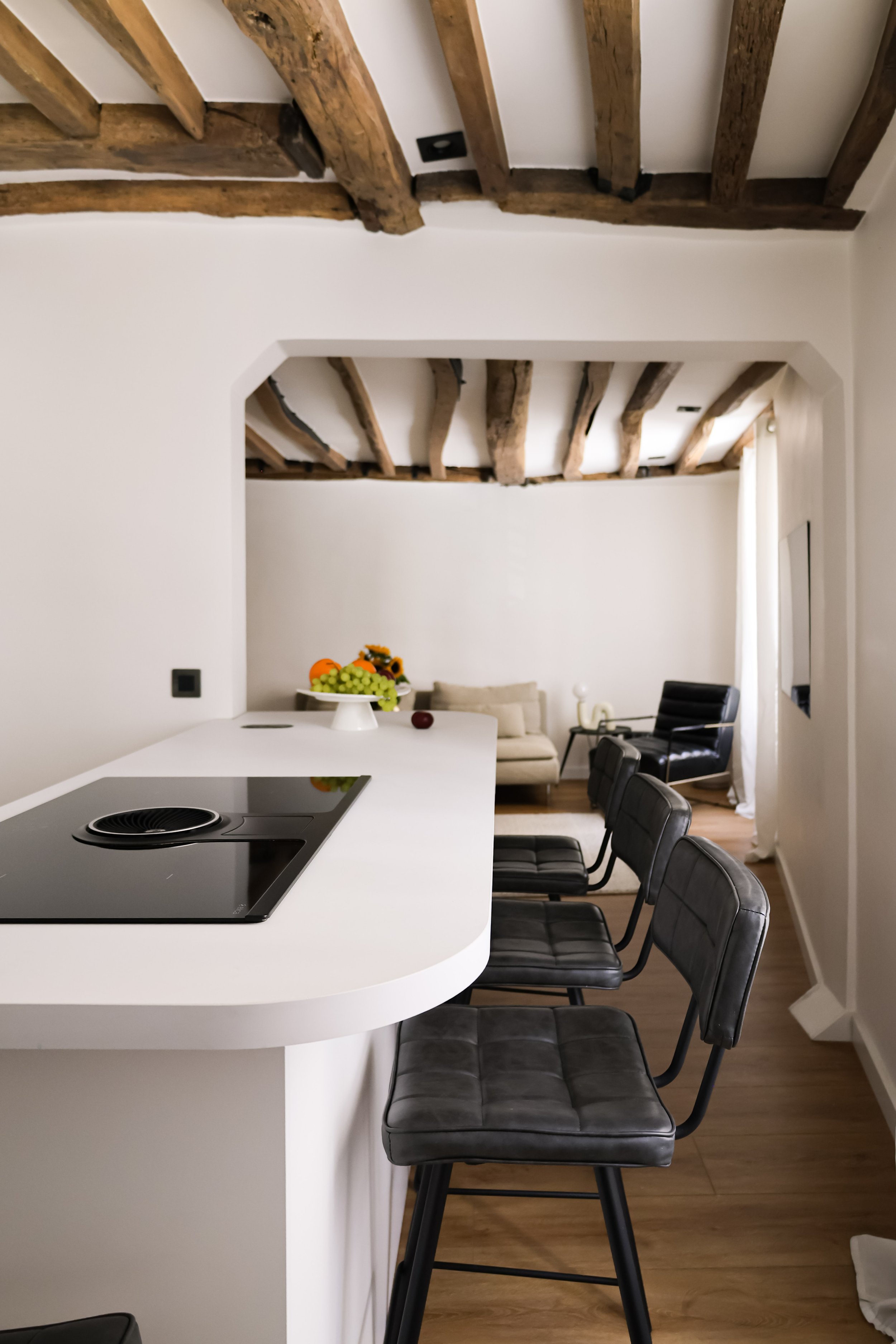 Intérieur d'une cuisine moderne avec îlot blanc, quatre chaises en cuir noir, meubles en bois clair, plafond avec poutres en bois apparentes et espace de vie en arrière-plan avec canapé beige et fauteuil noir.