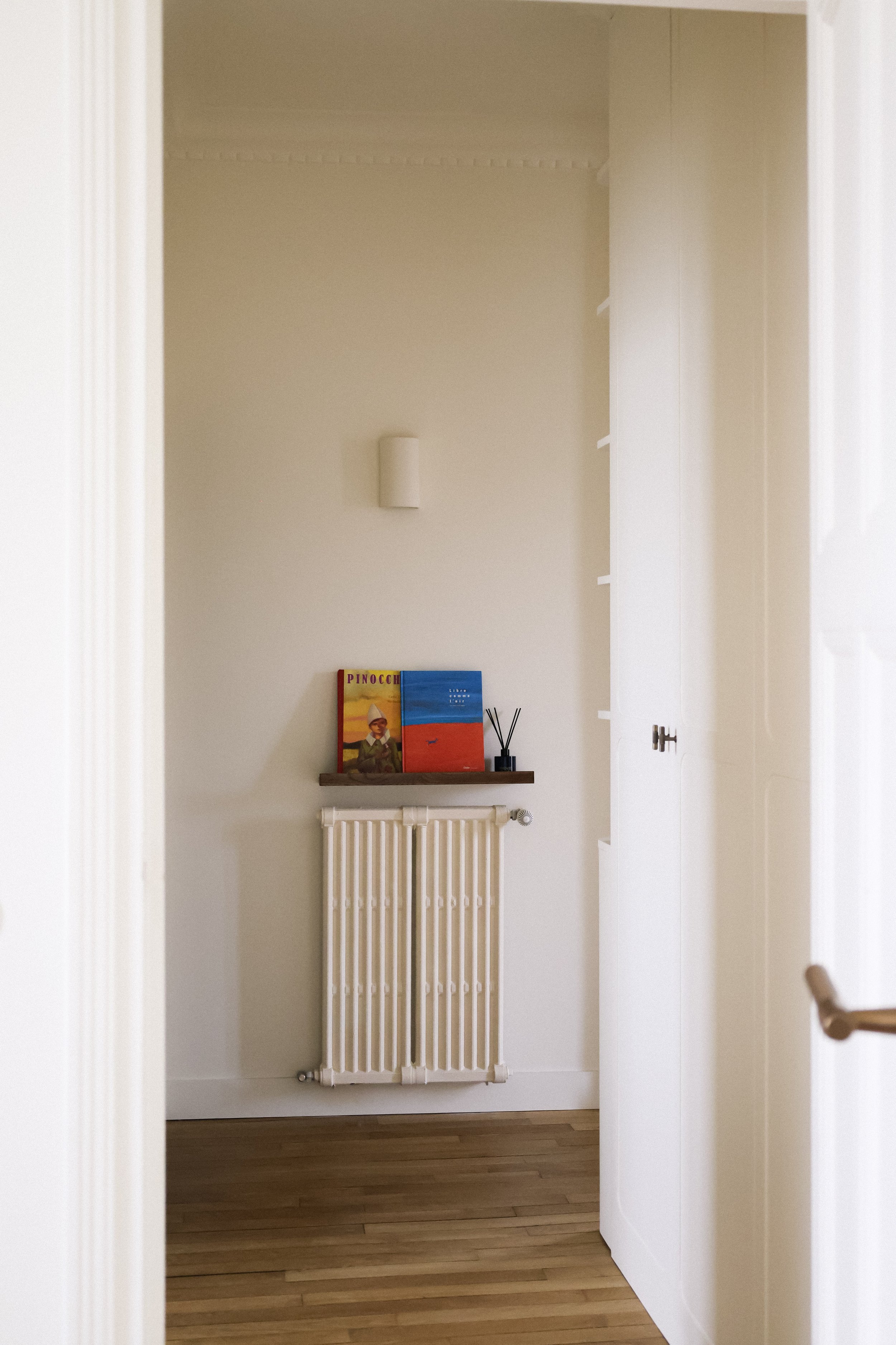 Vue d'un coin de pièce vue à travers une porte. On voit un radiateur blanc, une étagère en bois avec deux livres, un diffuseur d'encens et une petite lampe murale, le tout sur un mur blanc, avec un sol en bois.