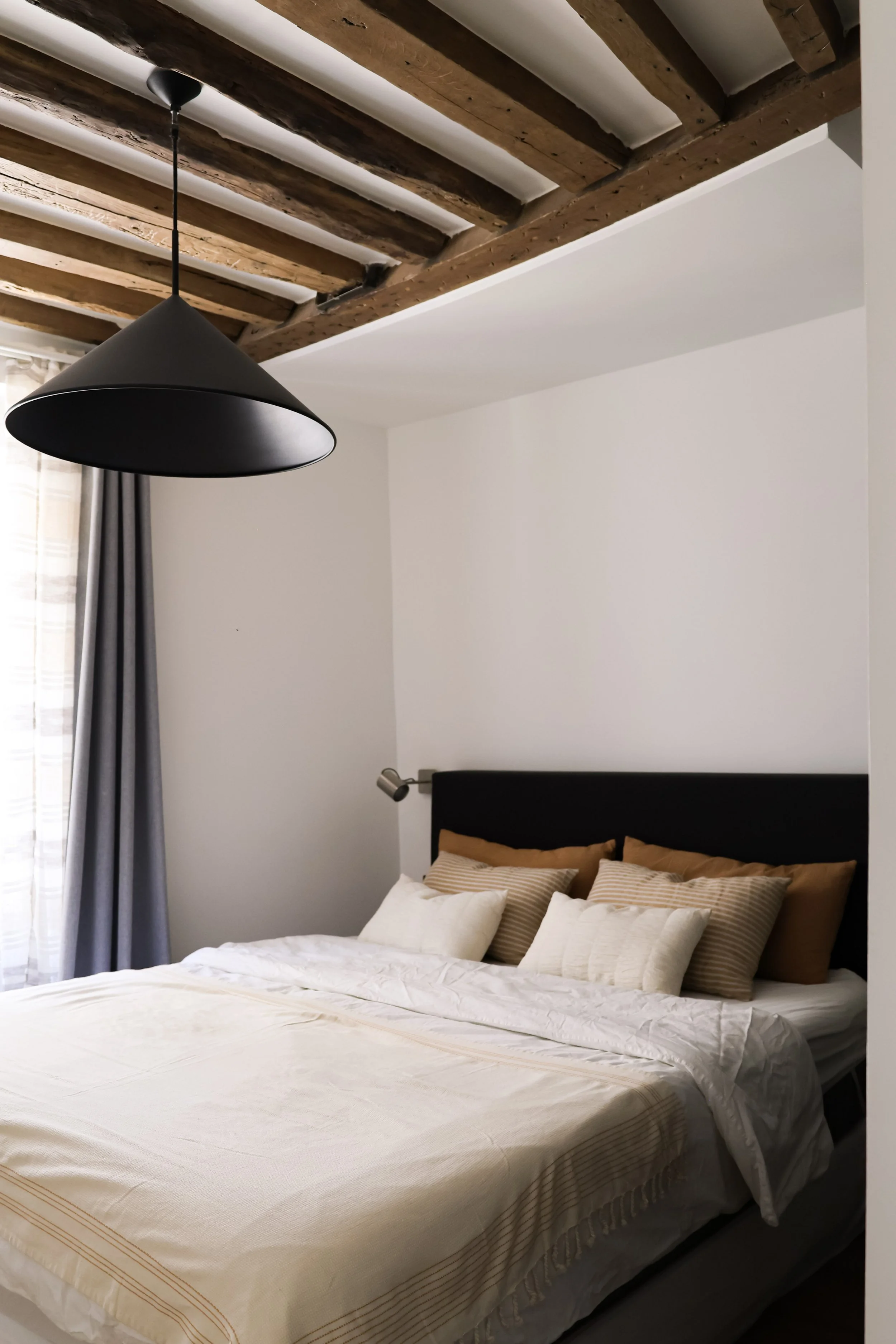Chambre à coucher minimaliste avec un lit, des coussins beige et blancs, un mur blanc, des rideaux gris, un plafond en bois et une suspension noire.