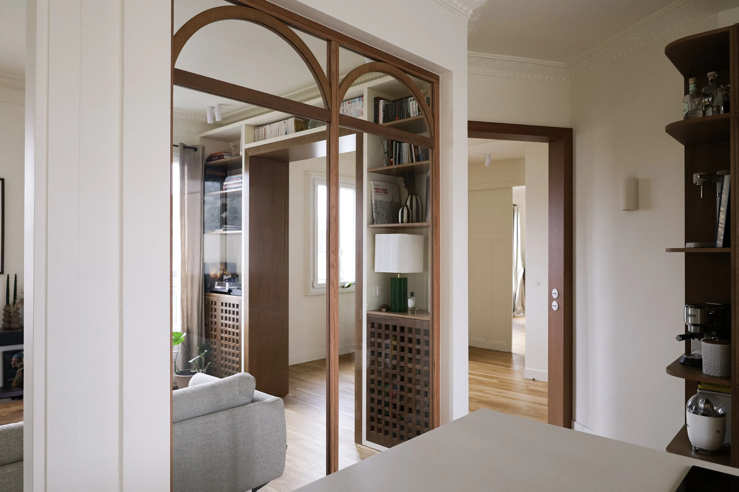 Intérieur d'un appartement avec une séparation en bois et miroir, un canapé gris, une bibliothèque, un lampadaire vert, une table à café et un sol en bois.
