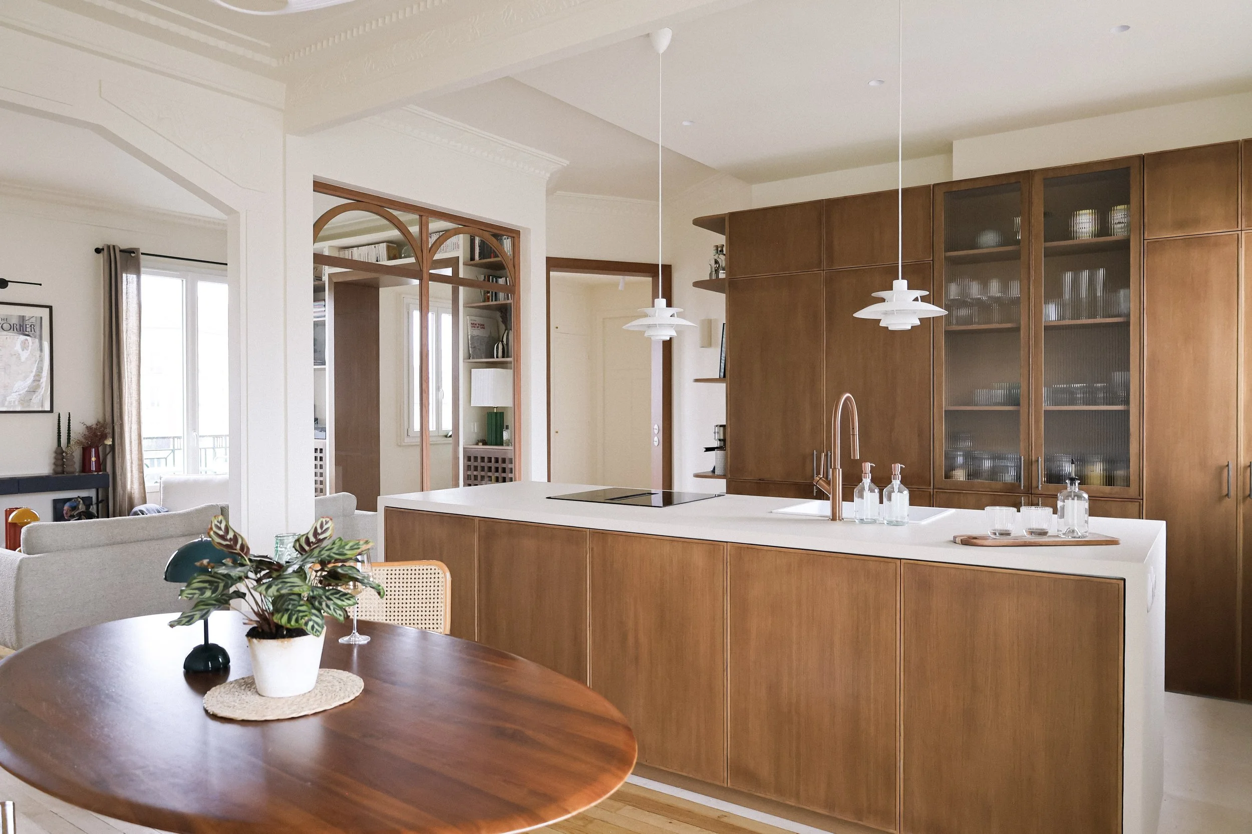 Cuisine ouverte avec une île en marbre blanc, armoires en bois, éclairage suspendu blanc, table ronde en bois avec vase de plantes