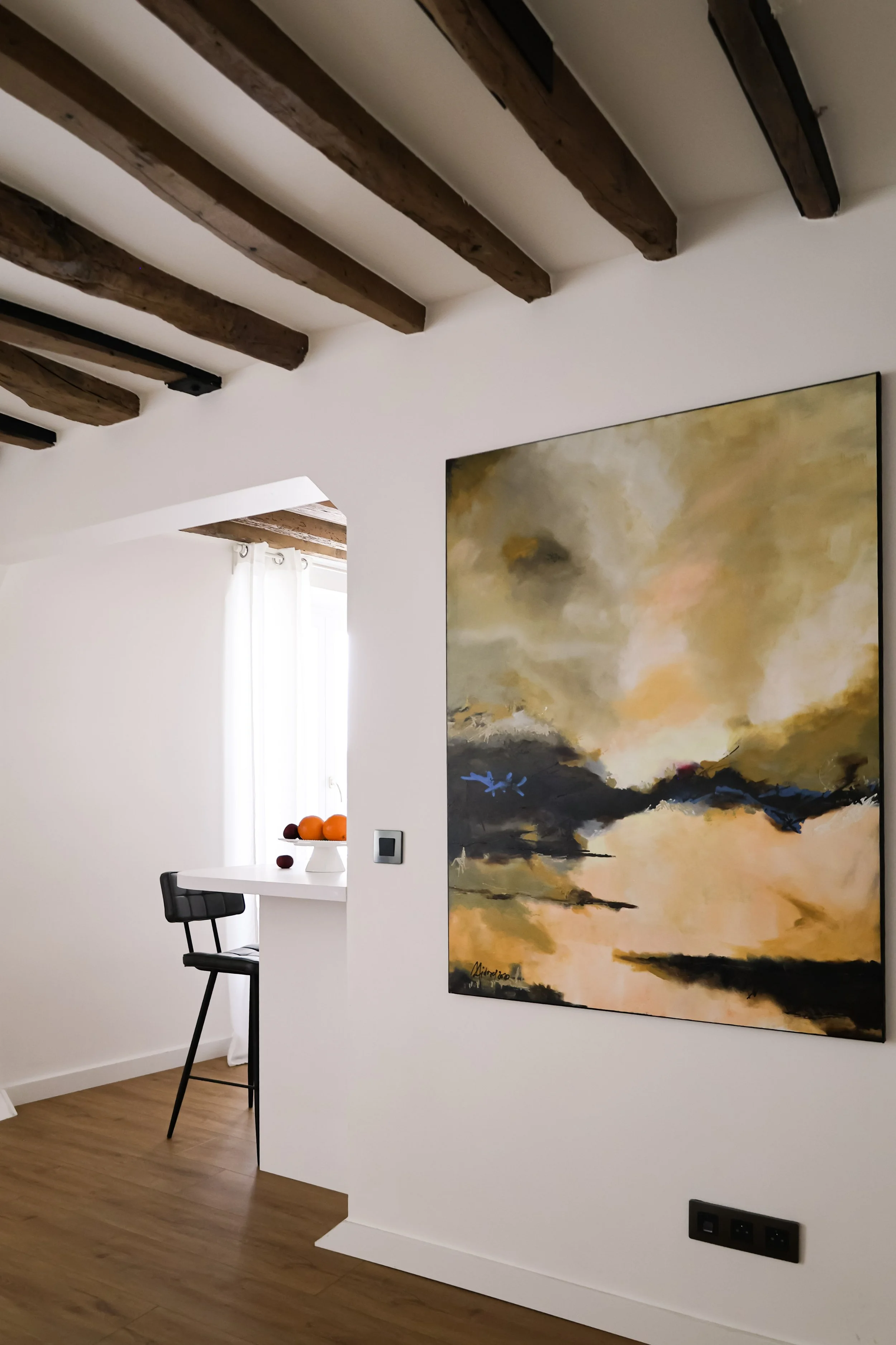 Une peinture abstraite avec des couleurs chaudes et des accents de bleu, accrochée sur un mur blanc dans un intérieur moderne. On voit un bar ou comptoir avec des oranges et un siège noir à côté d'une fenêtre avec des rideaux blancs.
