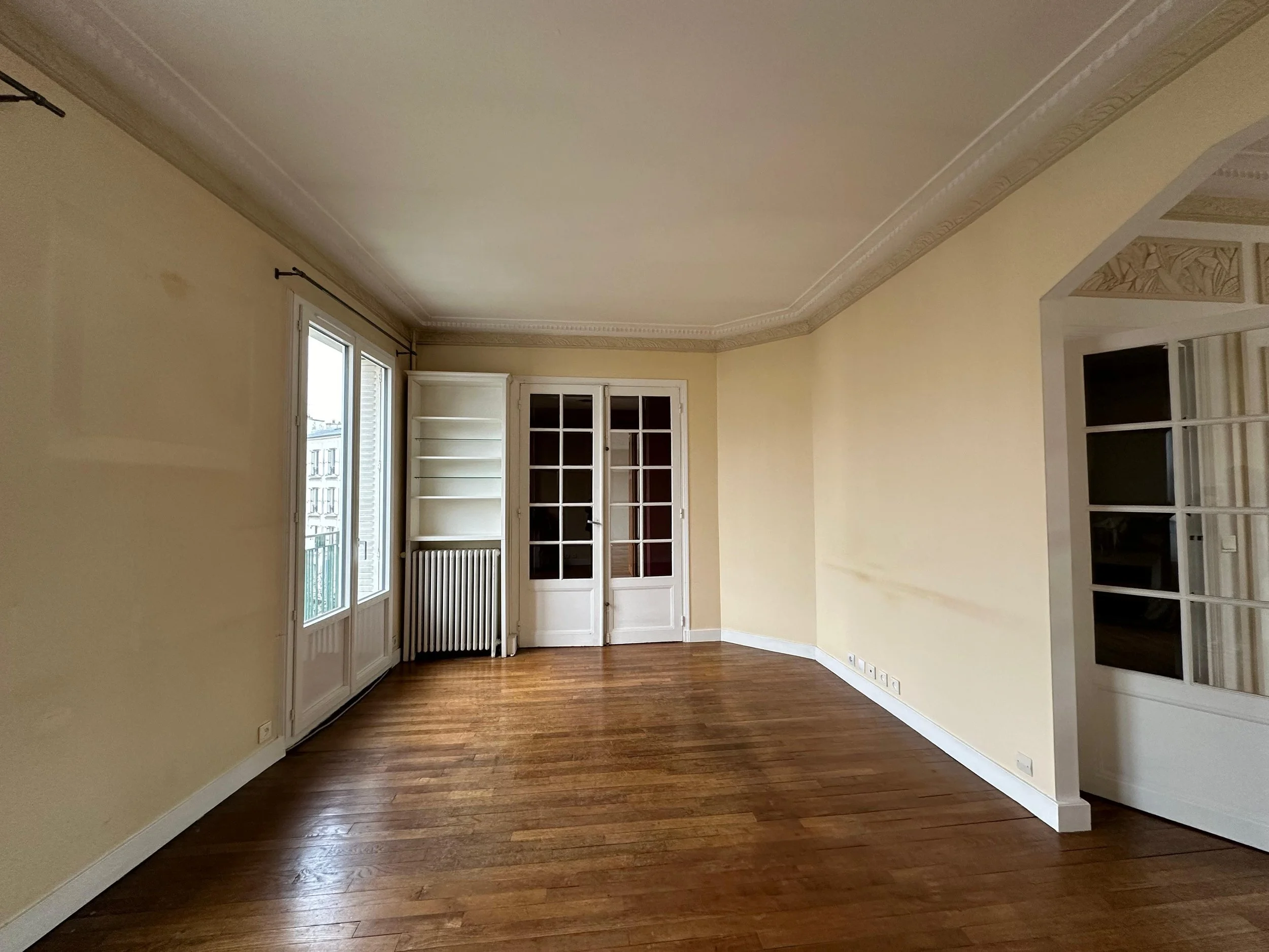 Salon vide avec fenêtres, parquet en bois, murs beige clair, porte-fenêtre à double porte, étagère et radiateur.