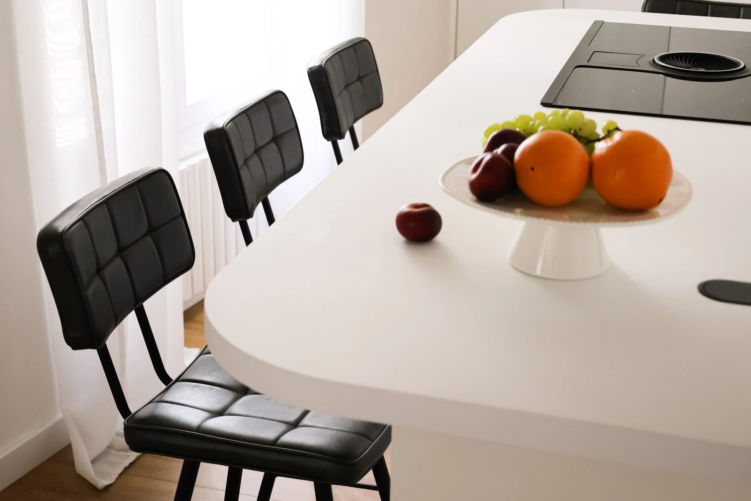 Une table de cuisine blanche avec trois chaises en arrière-plan. Sur la table, un plateau avec des fruits tels que des pommes, des oranges, du raisin vert et des raisins rouges.