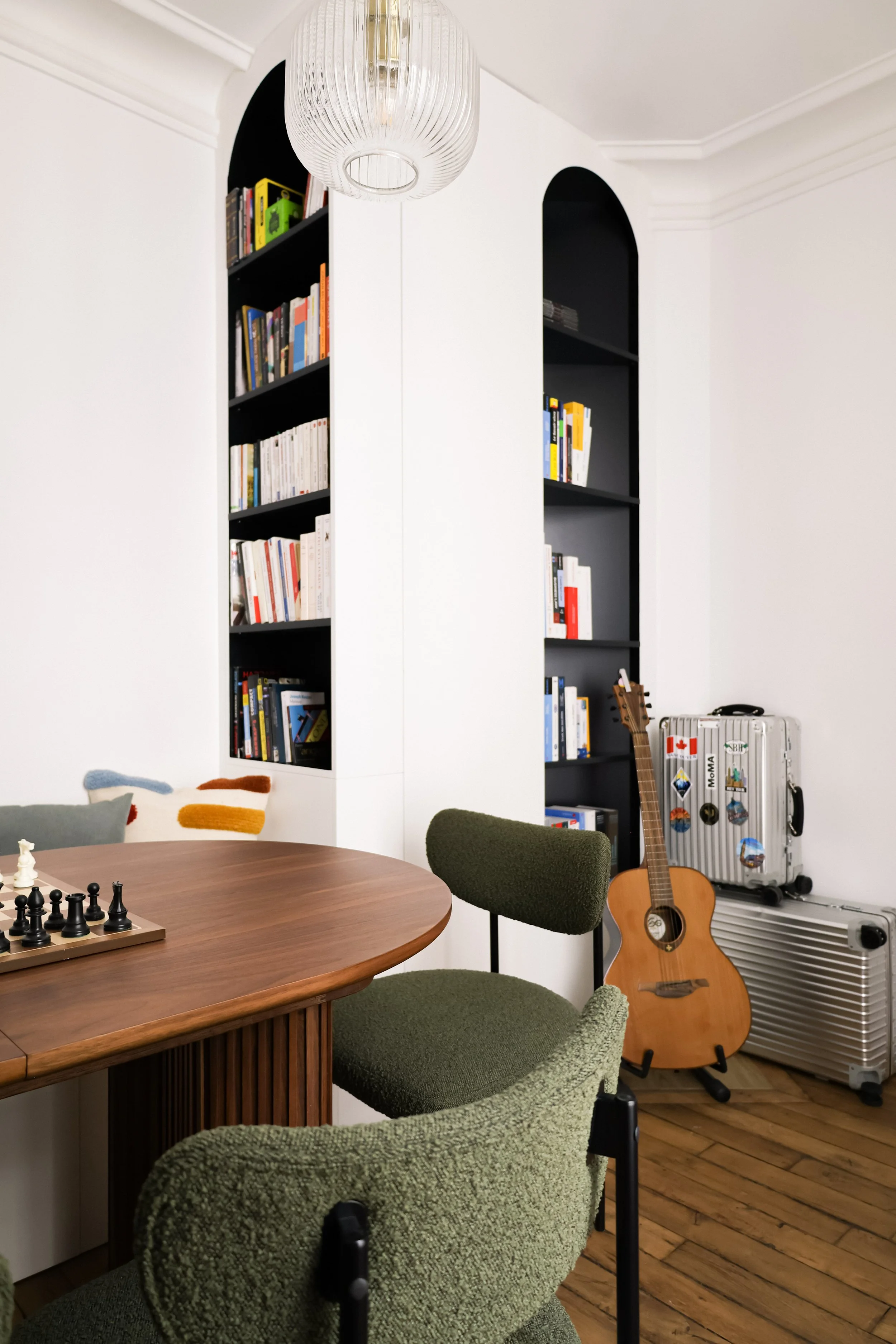 Salon avec un mur blanc, étagères noires remplies de livres, une guitare acoustique, deux valises, un fauteuil vert et une table ronde en bois.