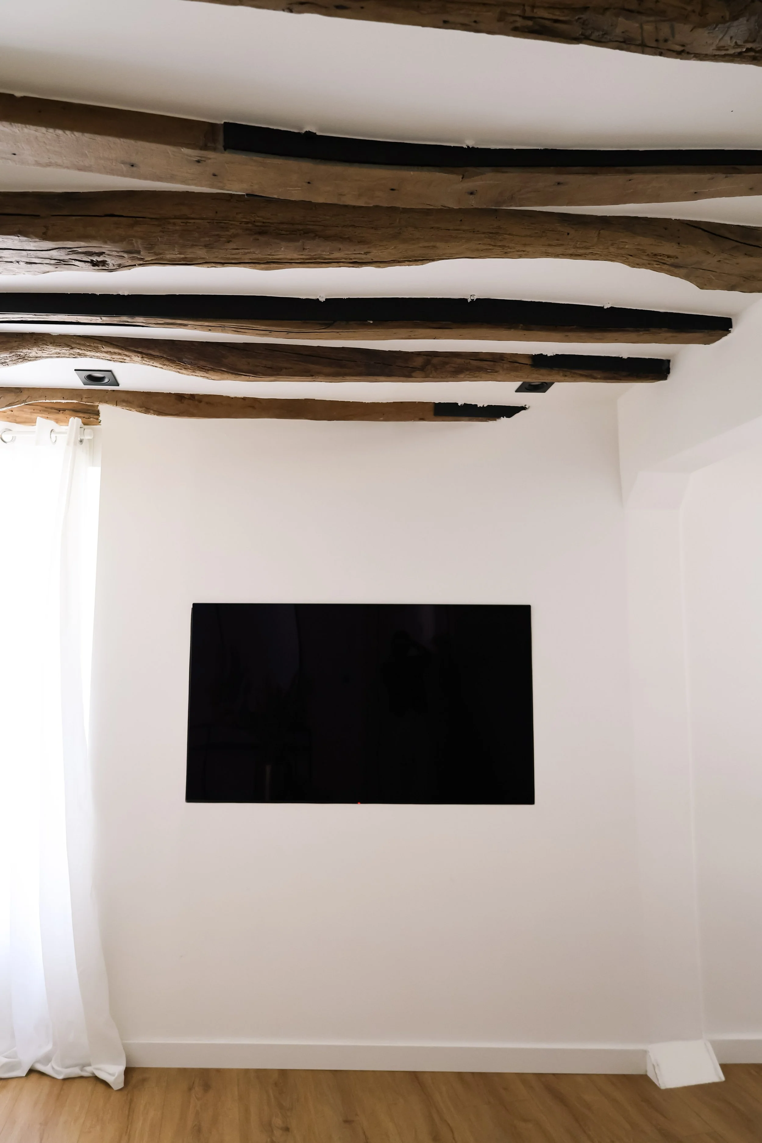 Mur blanc avec une télévision à écran plat noire, volet blanc, sol en bois, poutres en bois au plafond.