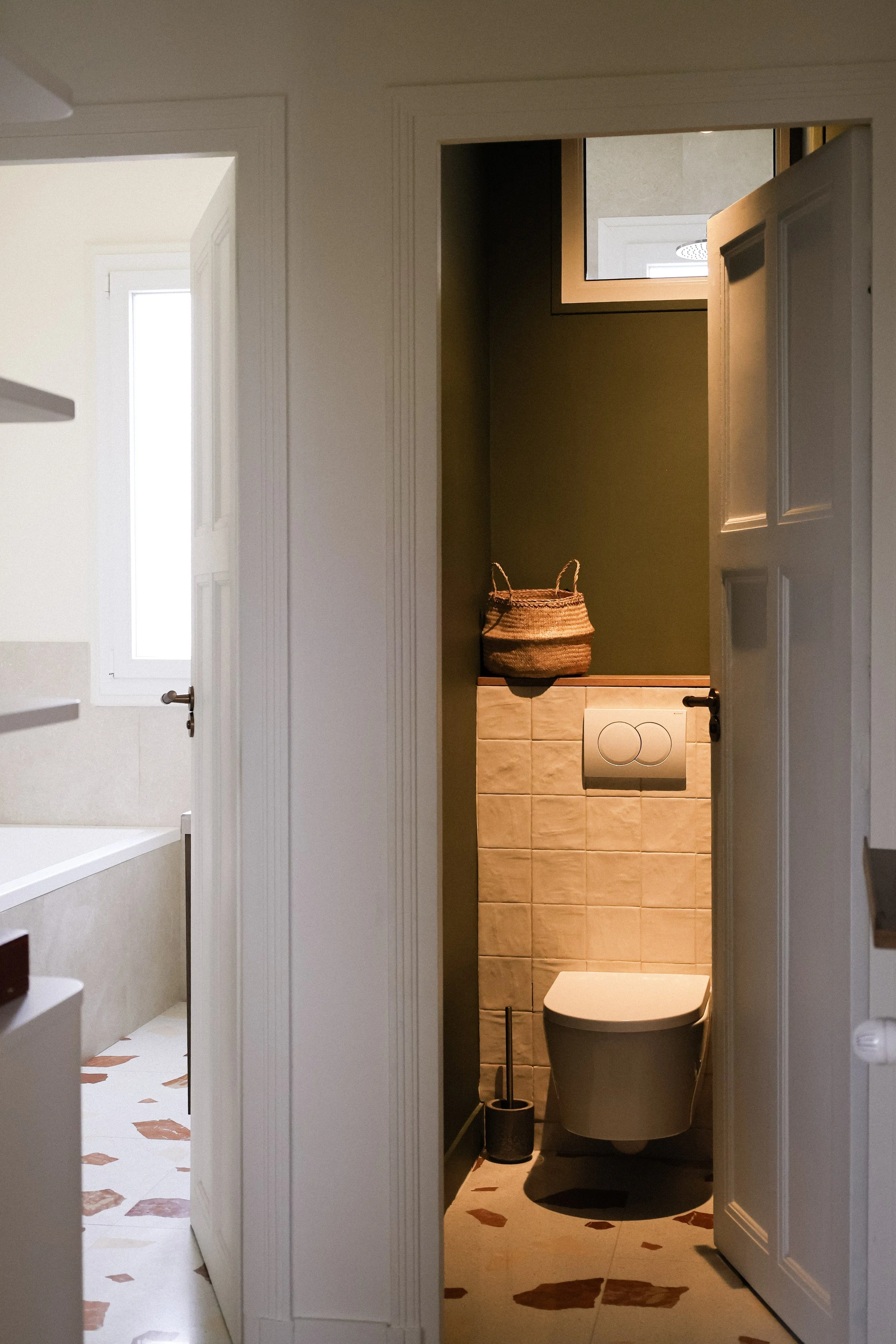 Petite salle de bains avec toilette suspendue, un panier en osier posé sur un mur peint en vert, et un petit fenêtre au plafond.
