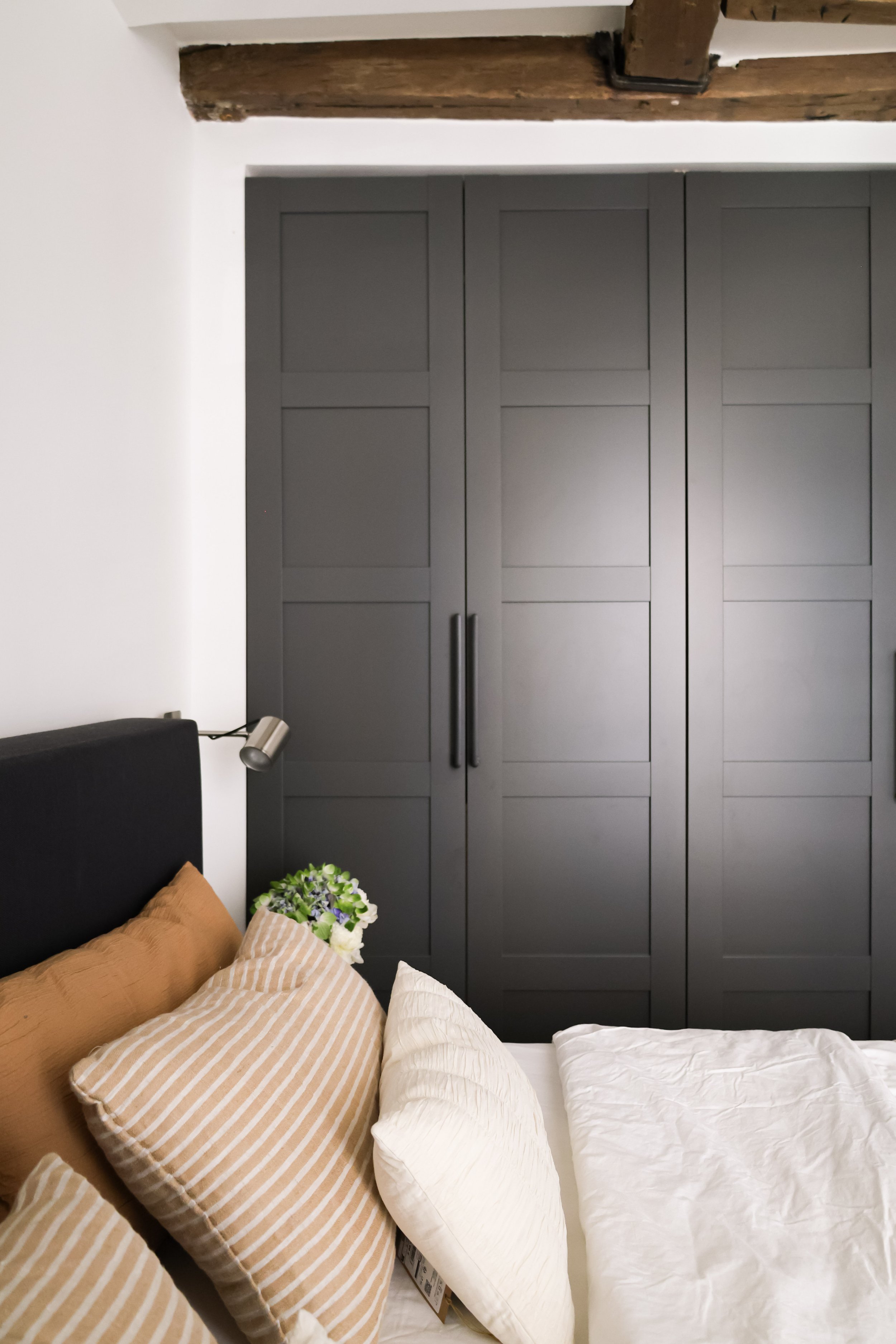 Chambre avec un lit, plusieurs coussins beiges et blancs, une plante et une armoire grise.