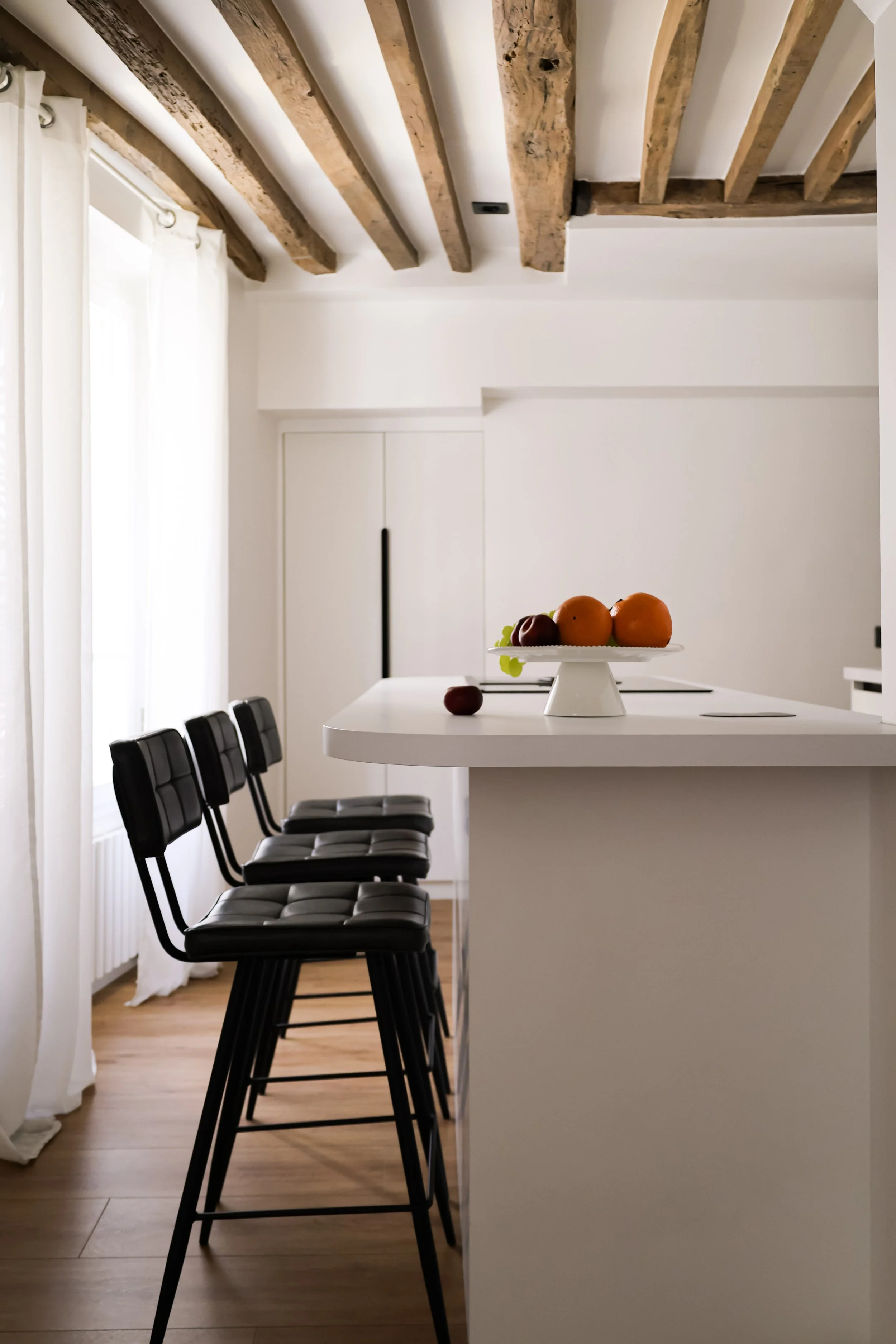 Cuisine moderne avec un plan de travail blanc, quatre chaises noires, un fruit sur un plateau blanc, poutres en bois au plafond, murs blancs et sol en bois.
