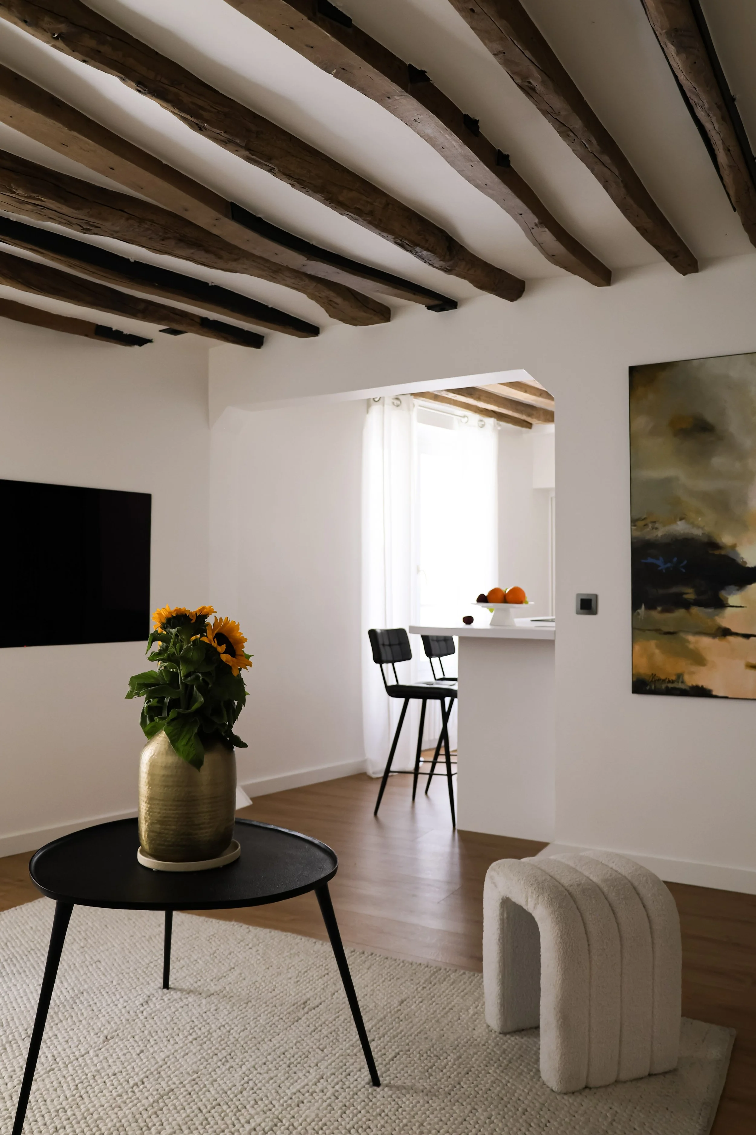 Salon intérieur avec plafond en poutres en bois visibles, une table basse noire avec un vase de tournesols, un tapis beige, une œuvre d'art abstraite sur le mur, et un canapé blanc.