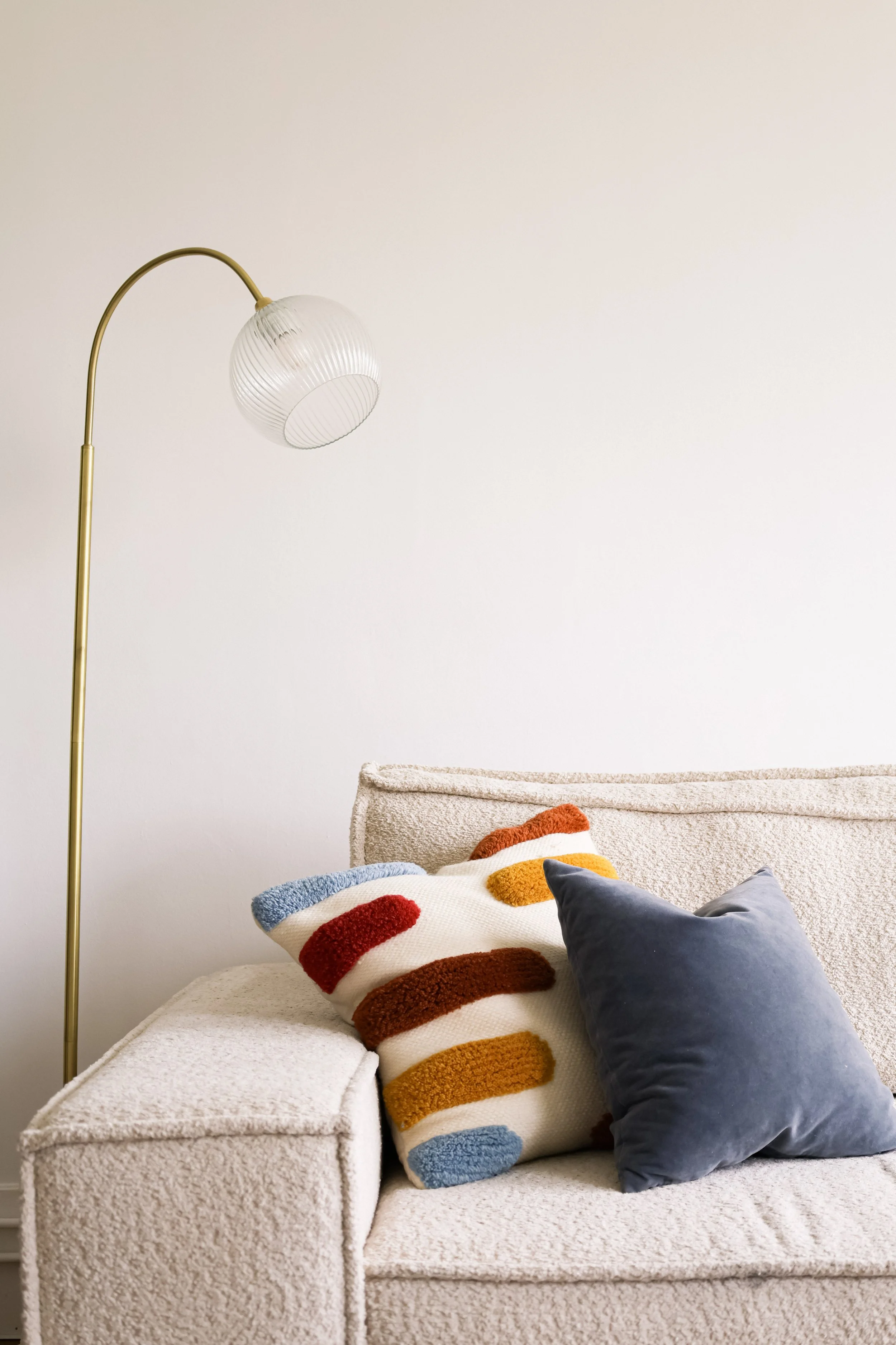 Canapé beige avec deux coussins, un à rayures multicolores en laine bouclée et l'autre en velours gris, et une lampe sur pied en or avec un abat-jour en verre. Mur blanc en arrière-plan.