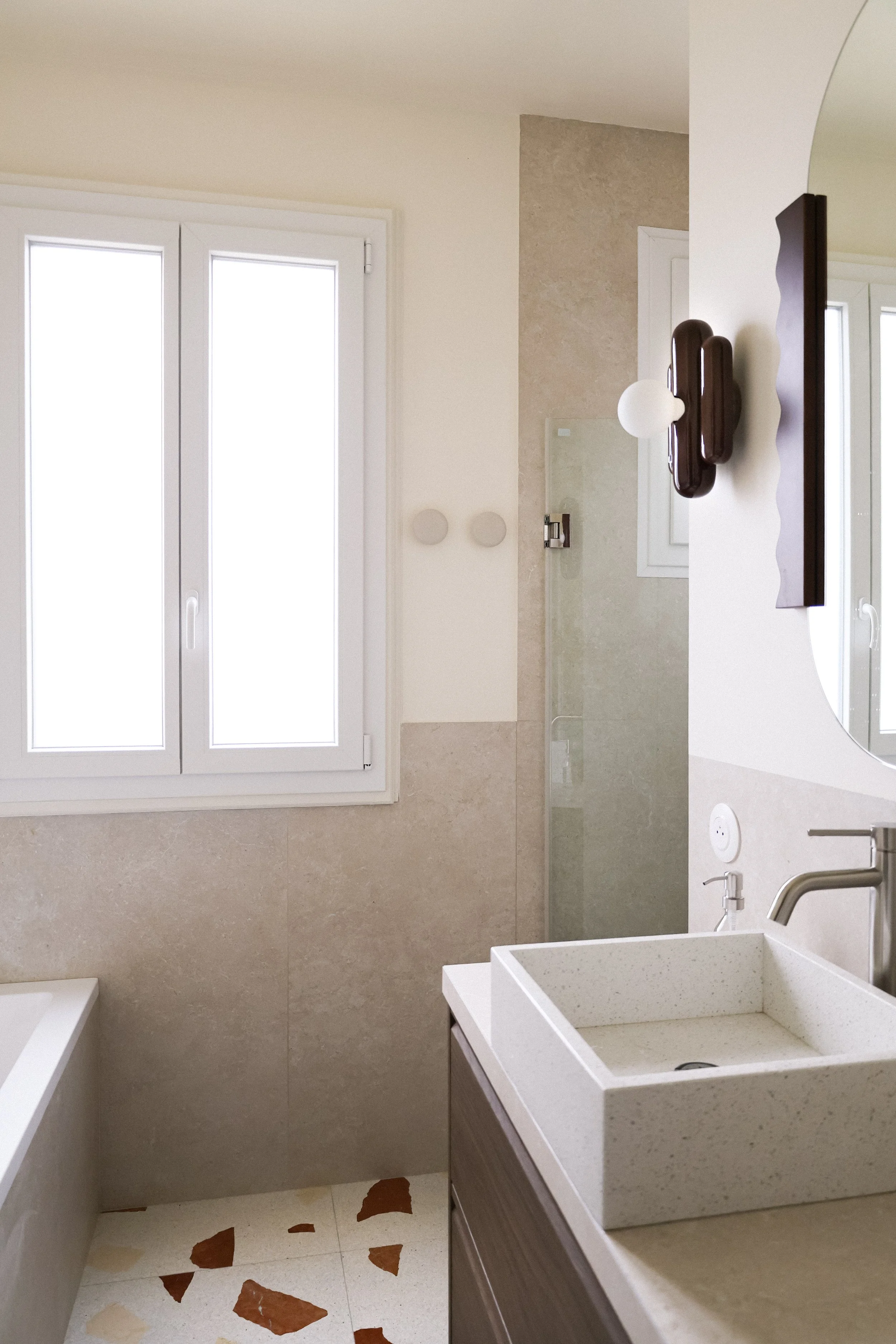Vue d'une salle de bain moderne avec une fenêtre, un lavabo carré, un miroir, une paroi de douche en verre, et un mur peint en blanc et beige