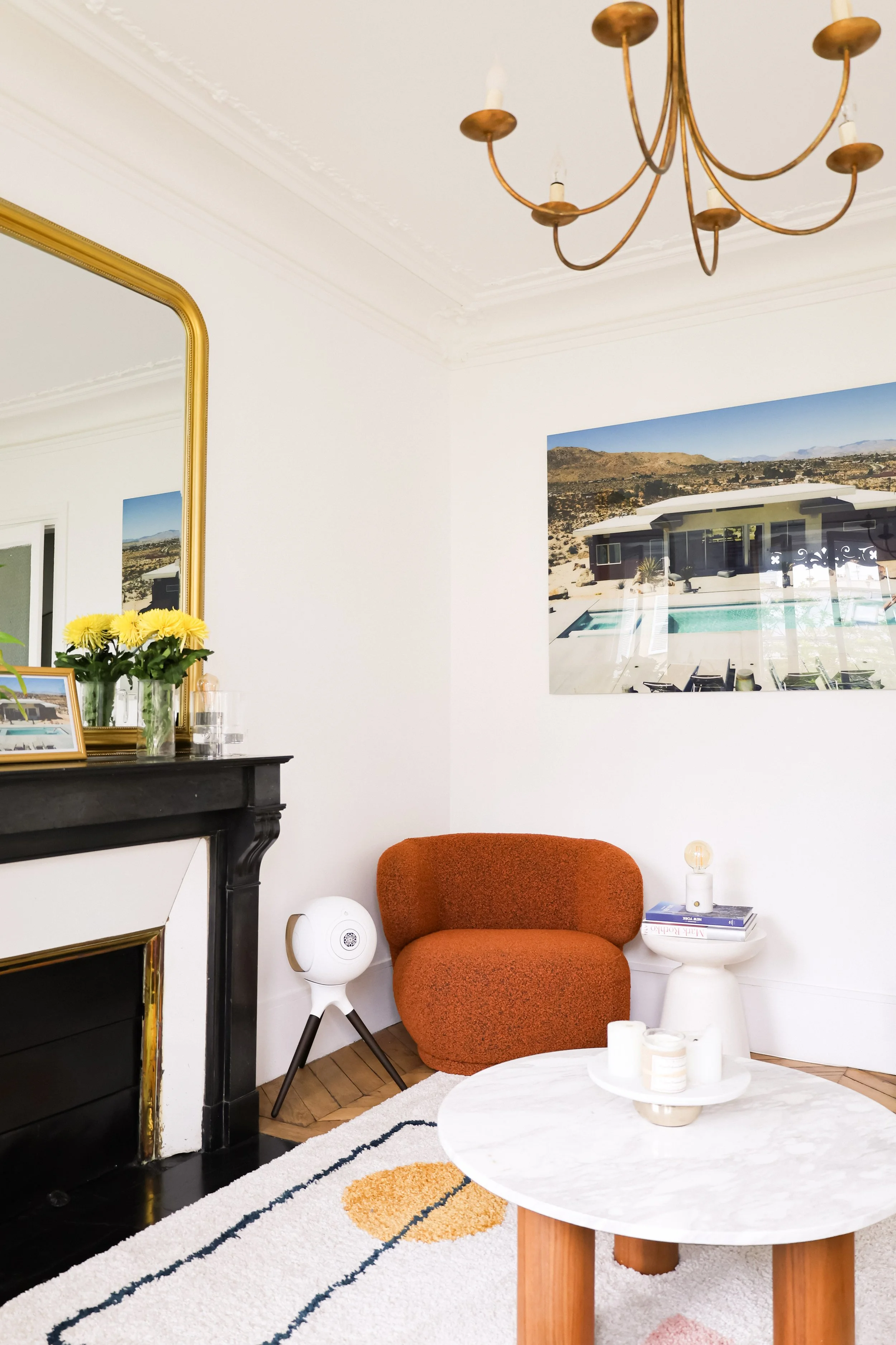 Salon moderne avec cheminée noire, miroir doré, tableau de paysage, fauteuil orange, petite table blanche avec livres et lampe, tapis blanc avec motifs, chandelier suspendu en laiton.