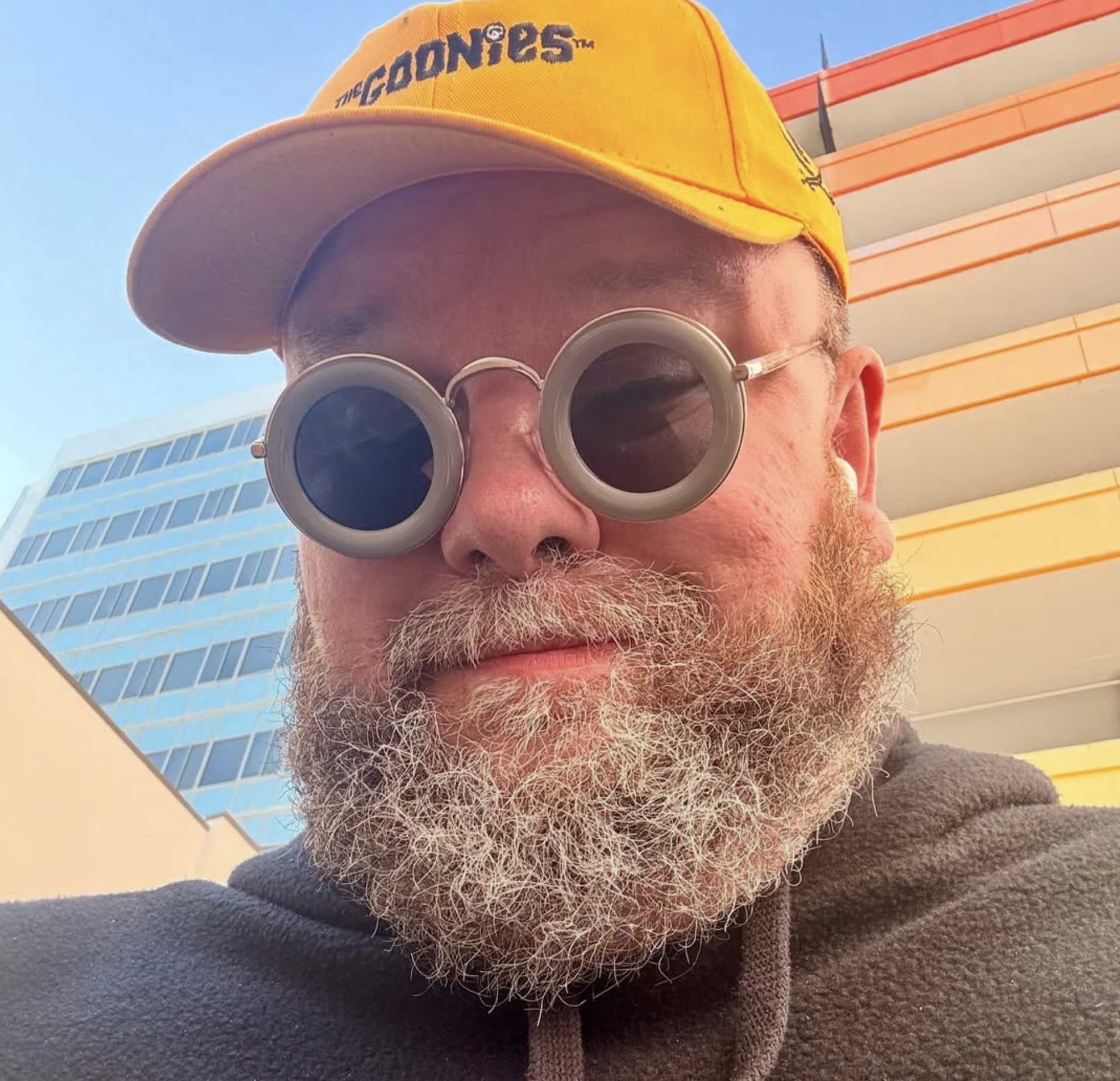 A man wearing a yellow The Goonies hat, round sunglasses, and a hoodie taking a selfie outdoors with modern buildings in the background.