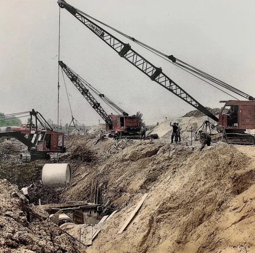 Construction site with large cranes and workers. Cranes are lifting large concrete pipes into a trench. Workers are guiding the placement of the pipes. The terrain is sandy with dirt mounds.