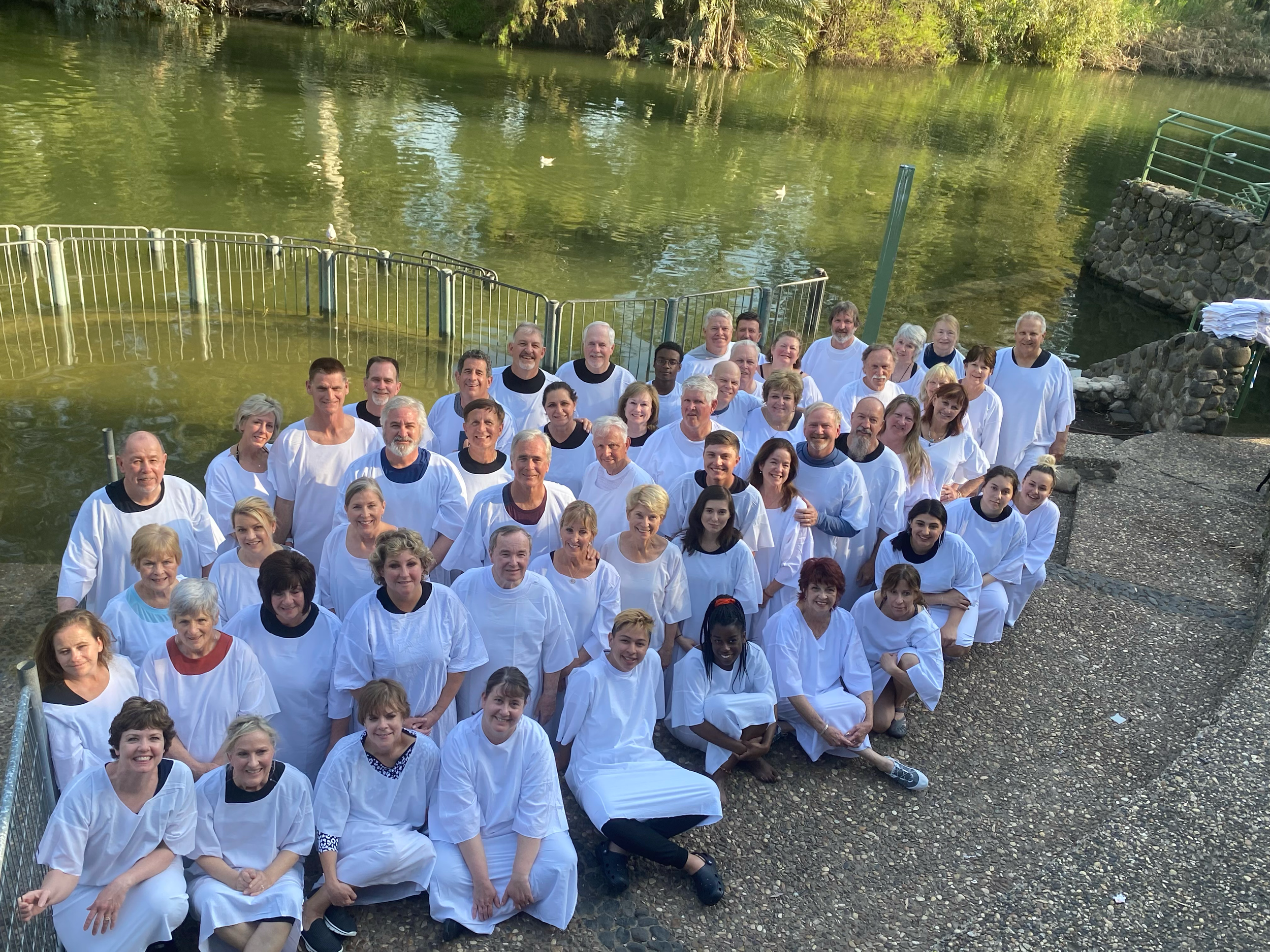 Group of people wearing white robes by a river, possibly for a baptism or religious ceremony.