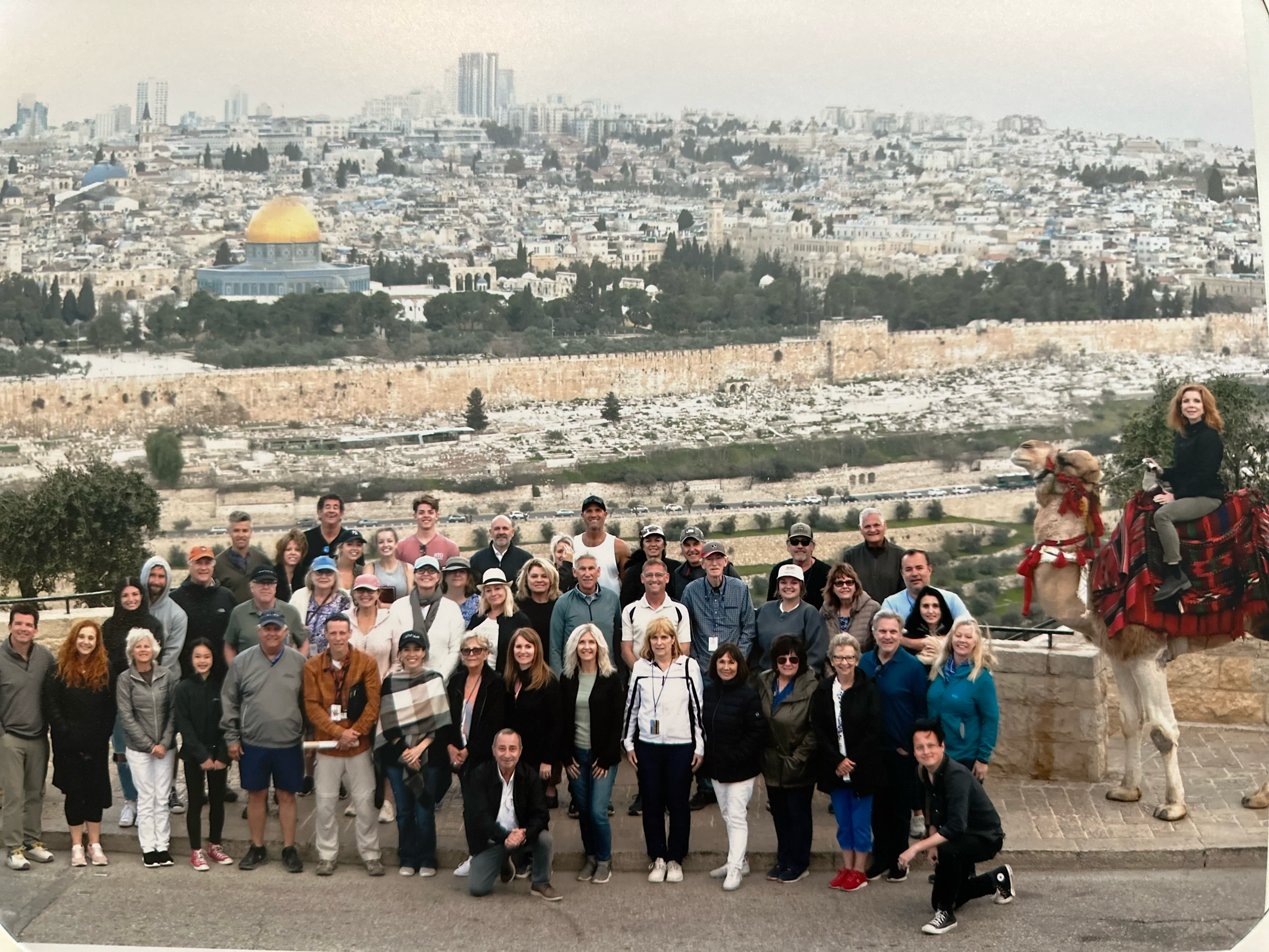 A group of people posing for a photo with a cityscape in the background, including a dome with gold features and a person riding a camel on the right.