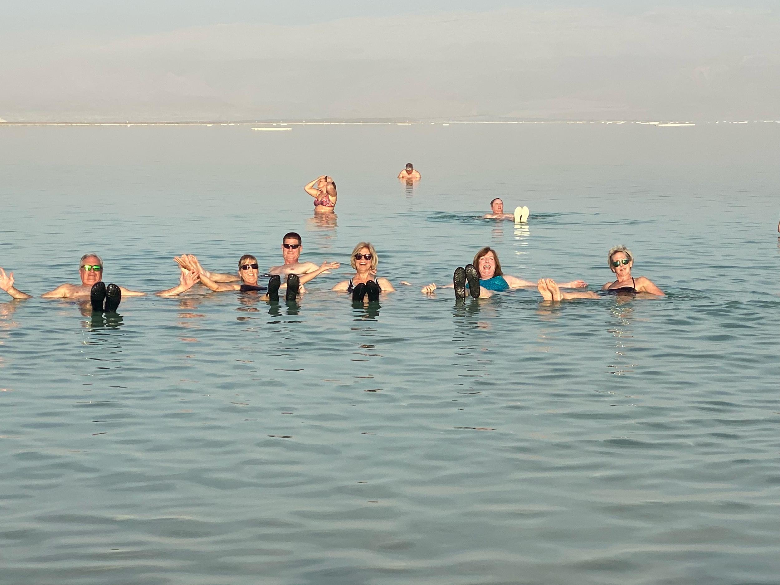 Group of people floating on their backs with raised feet in a calm body of water under a clear sky.