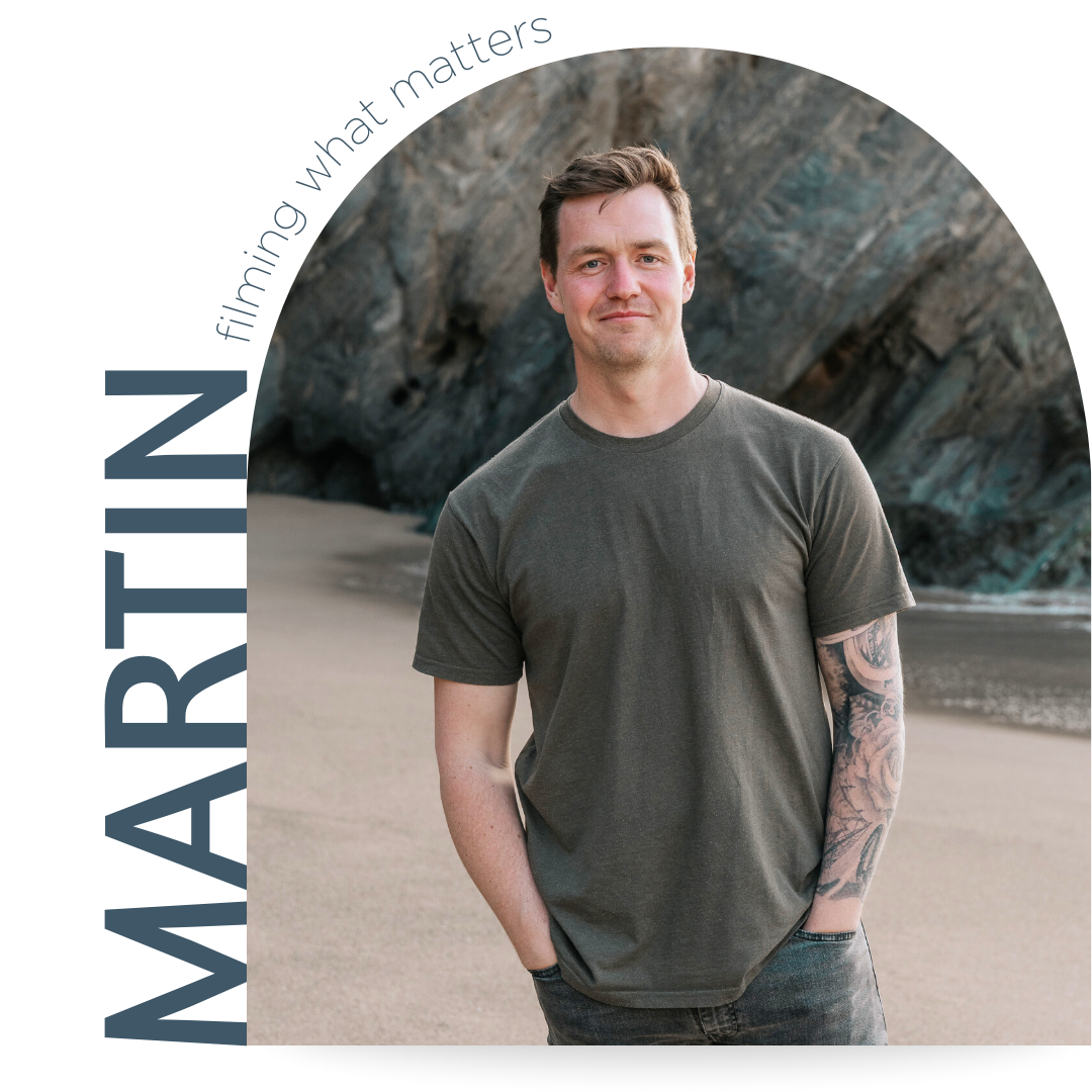 A man with short brown hair and a tattooed left arm stands on a beach with cliffs in the background, wearing a dark green t-shirt and jeans.