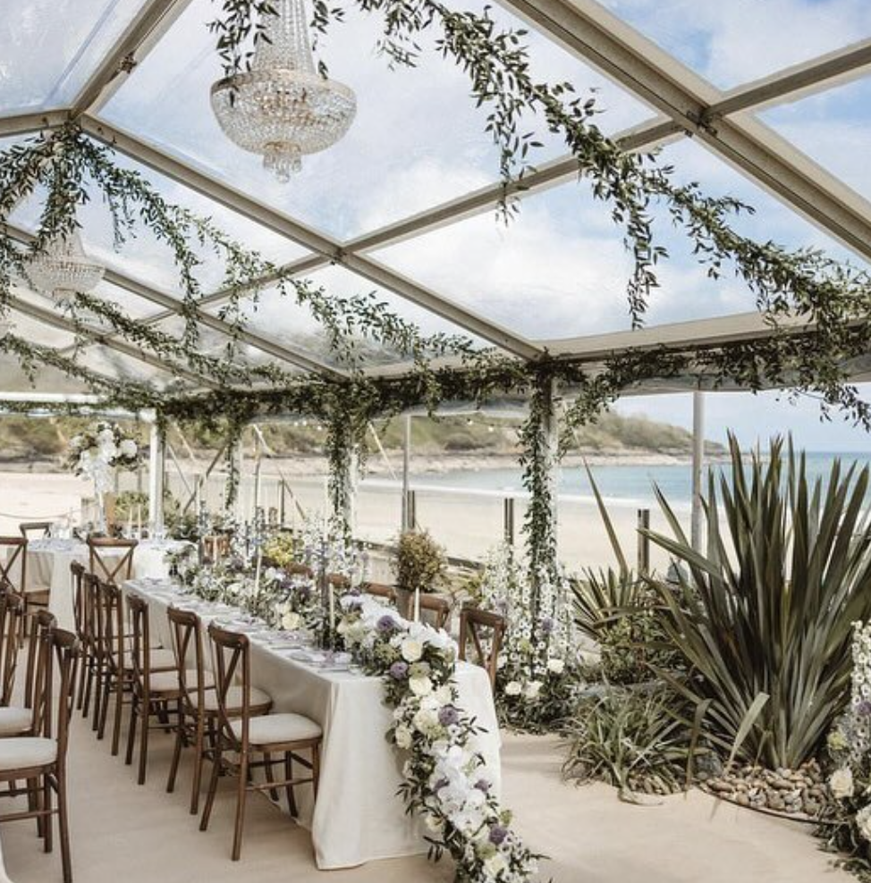 Elegant wedding reception setup under a glass canopy by the beach, decorated with greenery, chandeliers, and a long table with white floral arrangements.