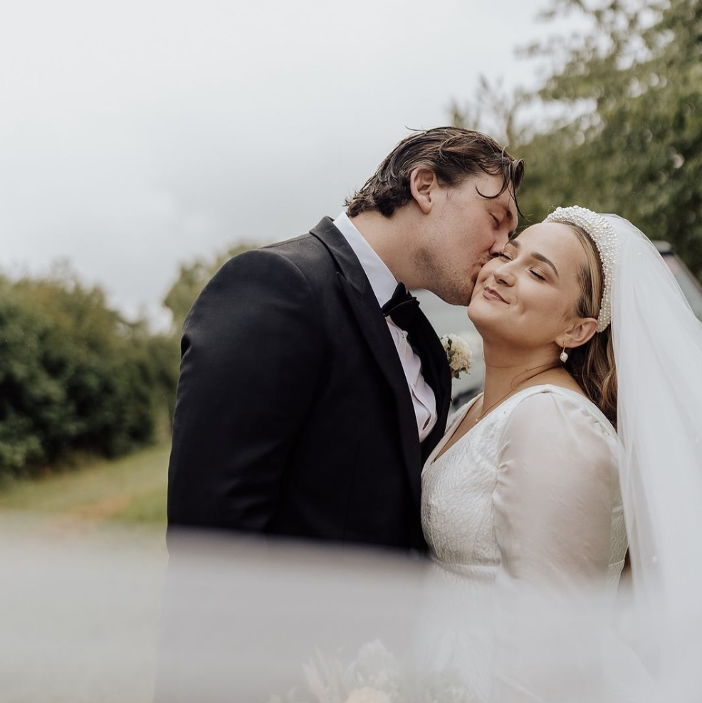 A groom in a black tuxedo kisses a bride wearing a white wedding dress and veil outdoors, with greenery in the background.